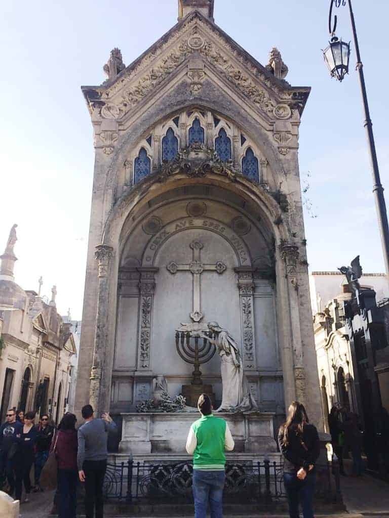 People stand in front of an ornate stone mausoleum adorned with religious symbols in a cemetery, discovering one of the unique things to do in Buenos Aires.