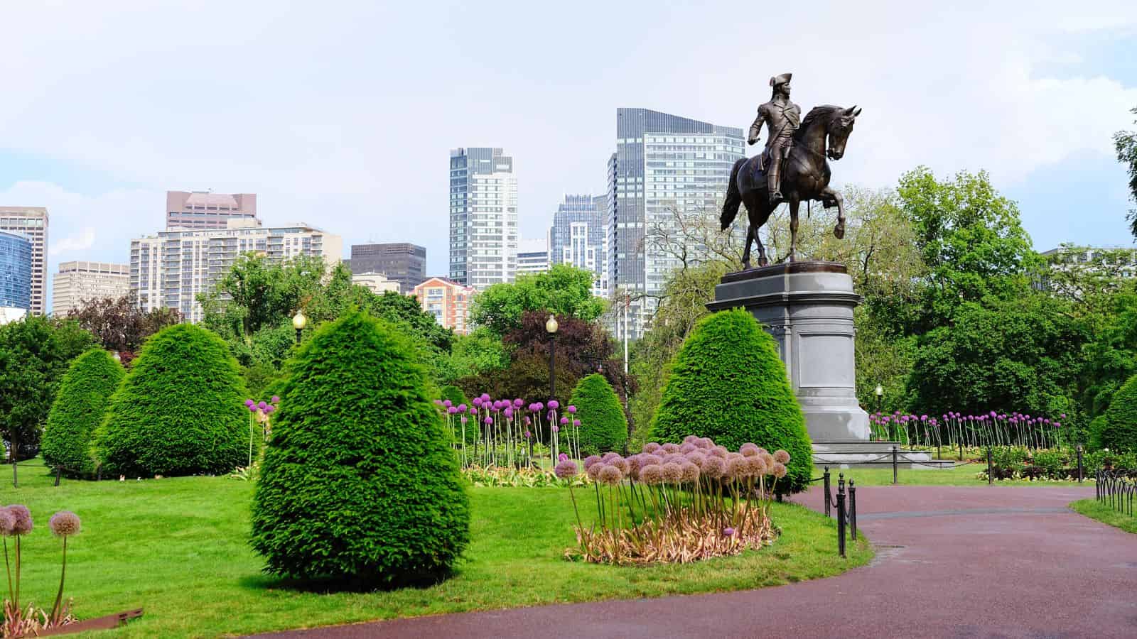Equestrian statue in a city park with green lawns, flowerbeds, and tall buildings in the background.
