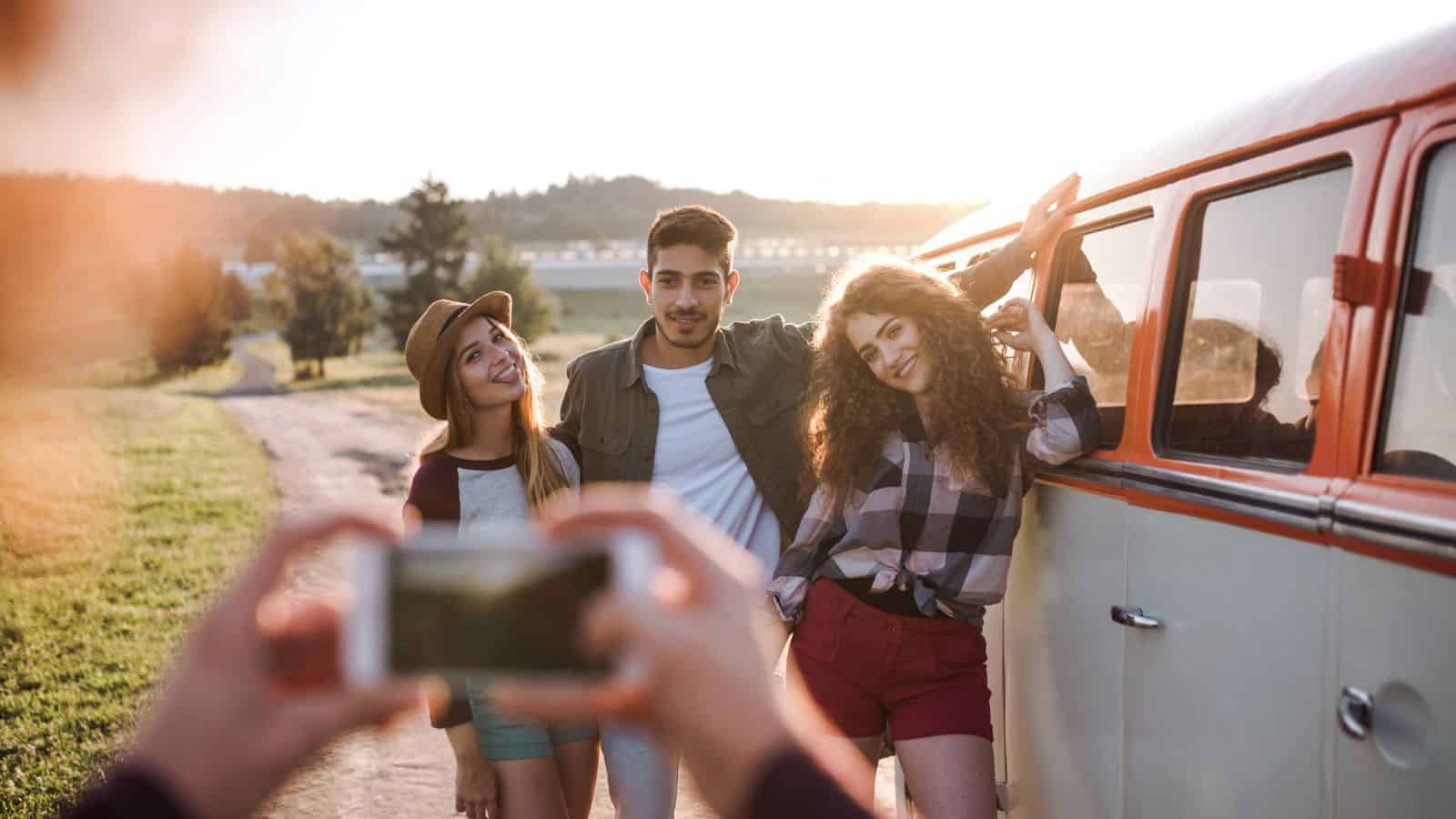 Three friends pose and smile beside a vintage van at sunset while someone takes their photo with a phone.
