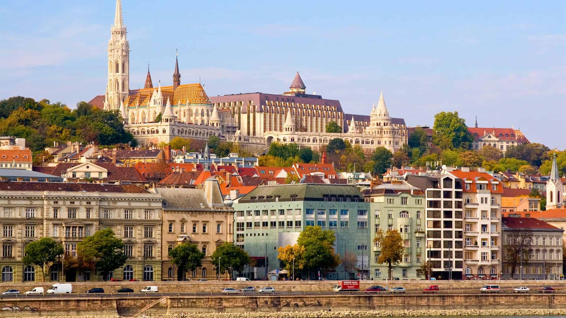 Colorful buildings and historic architecture along a riverbank in Budapest, Hungary, with a hilltop church.