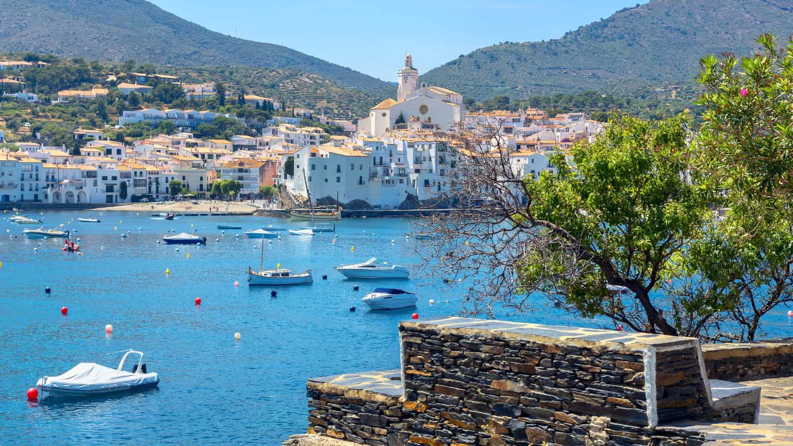 Boats float on blue water with a coastal town and hills in the background, viewed from a stone terrace.