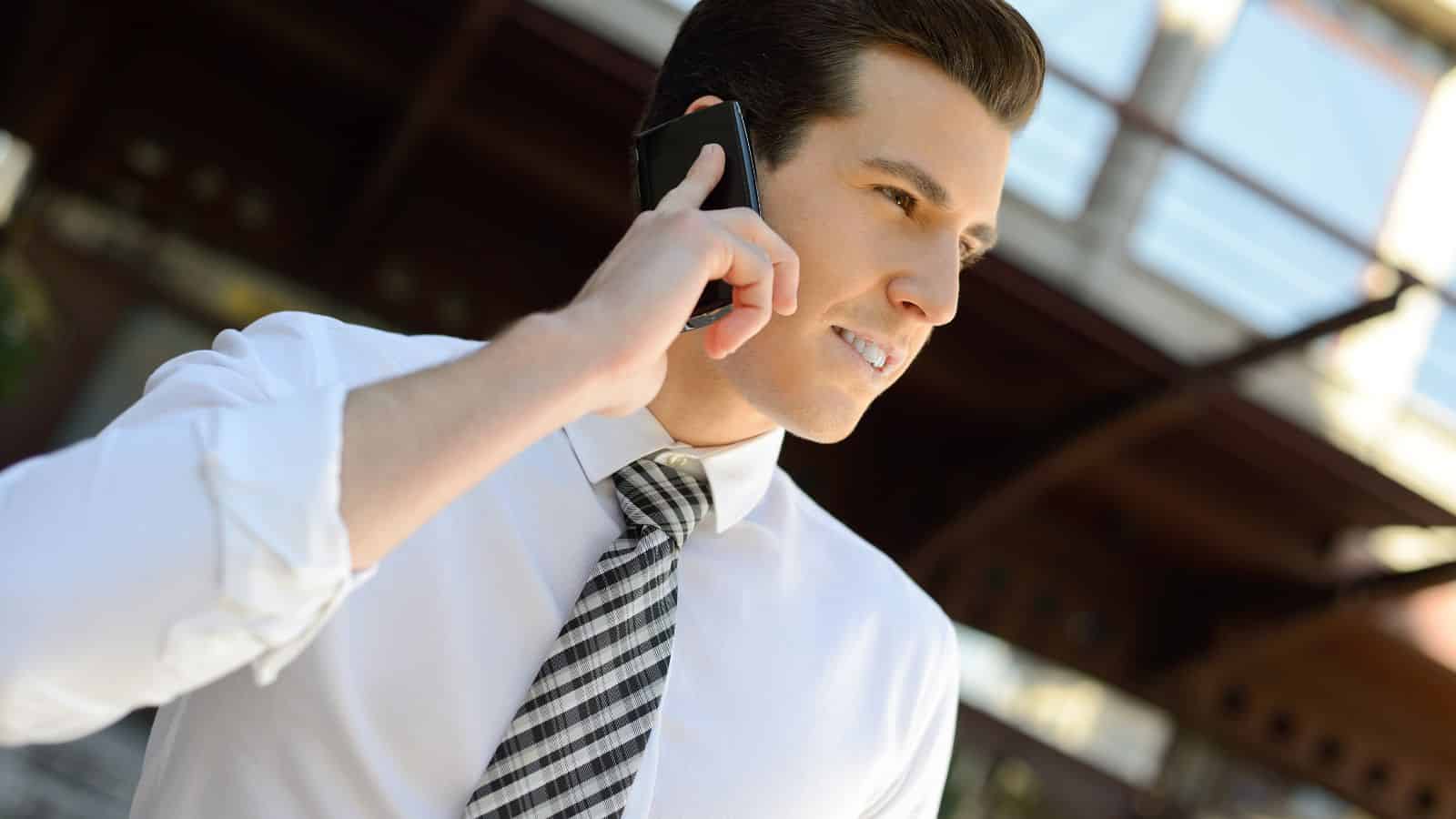 Man in a white shirt and plaid tie talking on a cellphone outdoors, looking concerned.