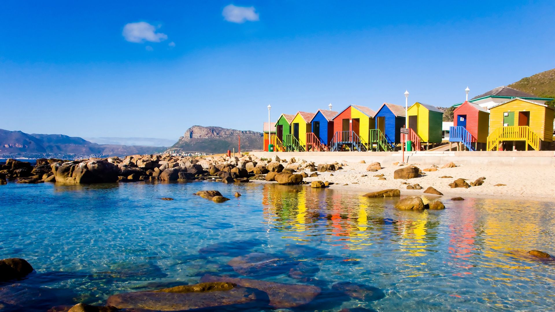 Colorful beach huts line a sandy shore with clear blue water and mountains in the background.