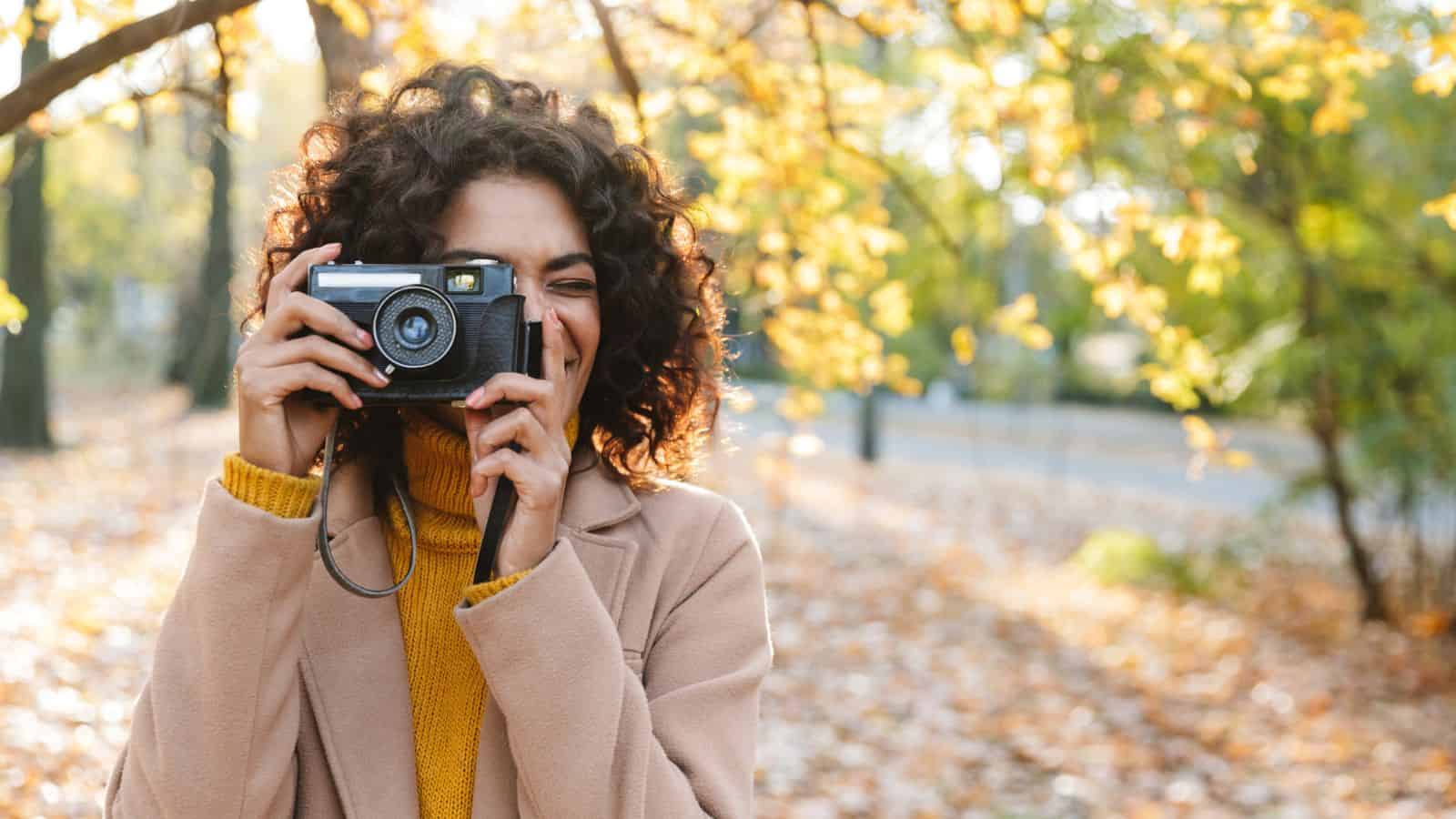 Woman with curly hair and a tan coat taking a photo outdoors in a park with autumn leaves.