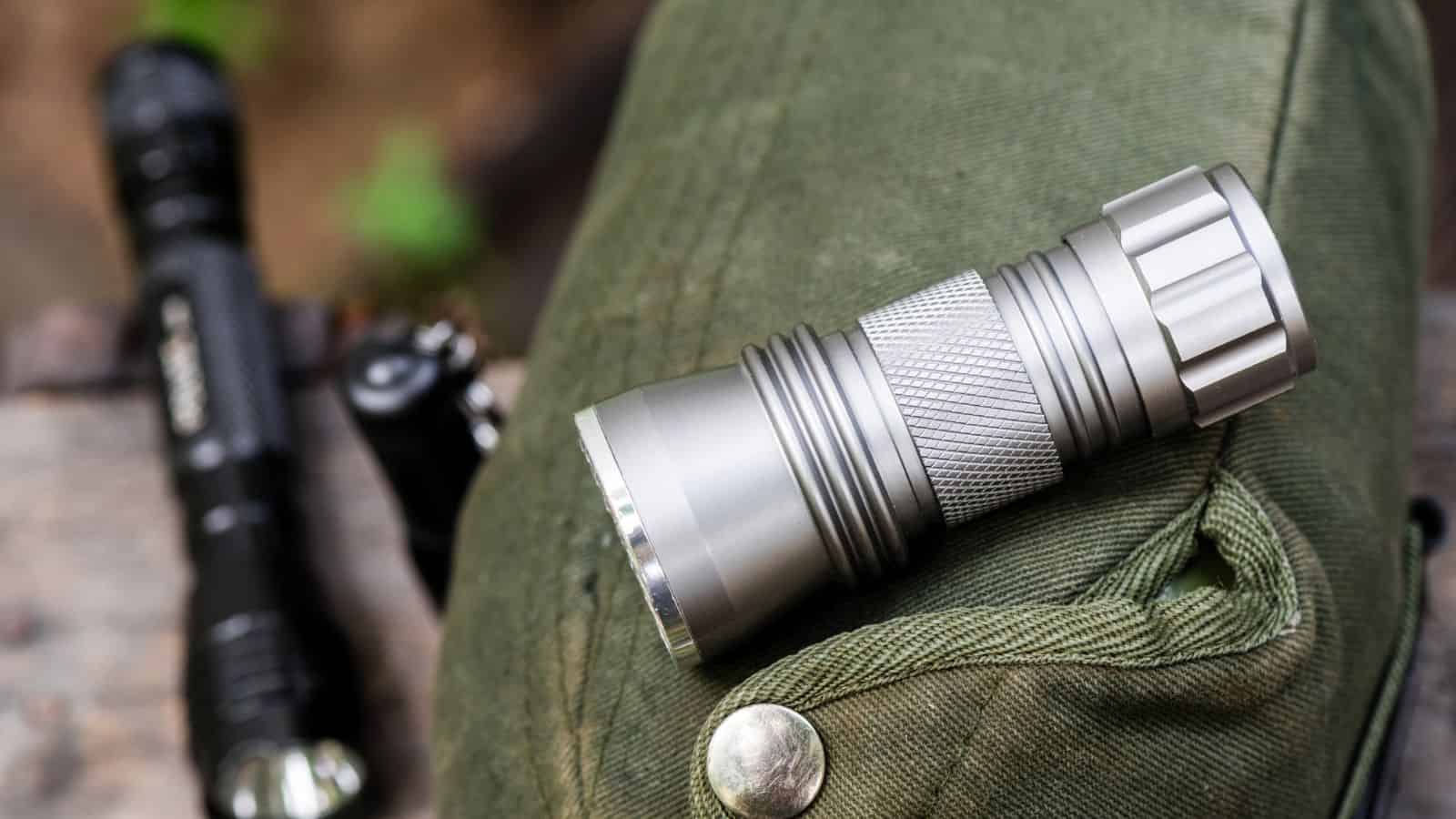 A silver flashlight resting on a green fabric bag, with a black flashlight blurred in the background.