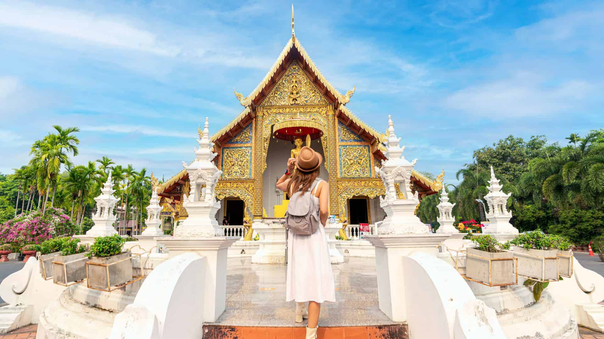 Woman in a hat stands in front of a golden temple with ornate details and lush greenery under a blue sky.