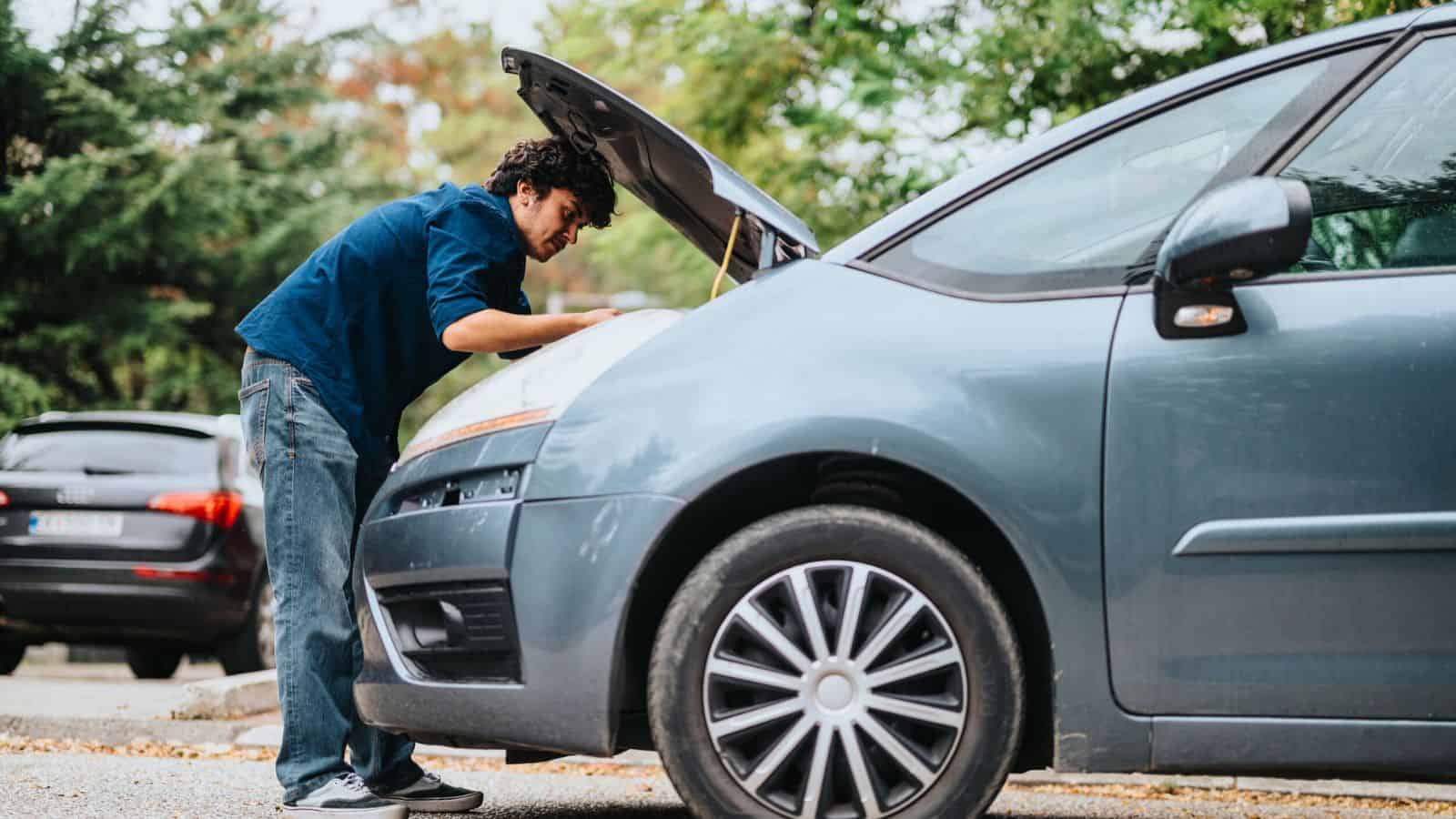 Man checking under the hood of a parked car on a street with trees and other vehicles in the background.