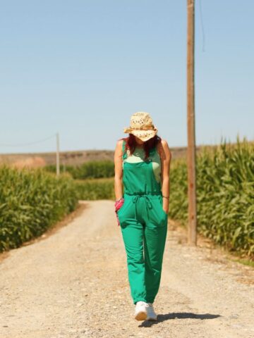 Woman in a green jumpsuit and hat walks down a dirt road between tall cornfields under a clear sky.