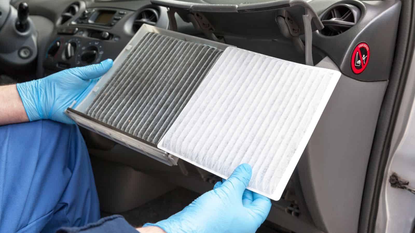 Gloved hands hold a dirty and a clean cabin air filter inside a car with the glovebox open.