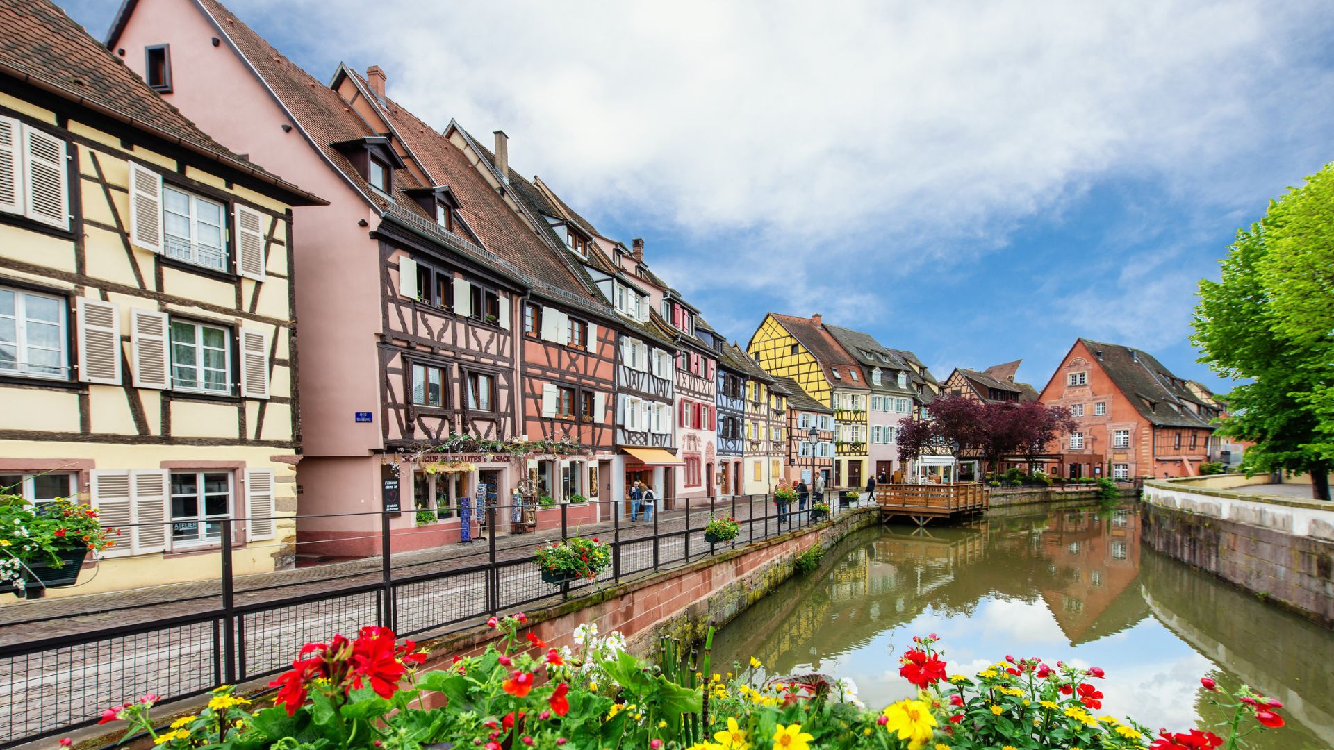 Colorful half-timbered houses line a canal with flowers in the foreground under a blue sky.