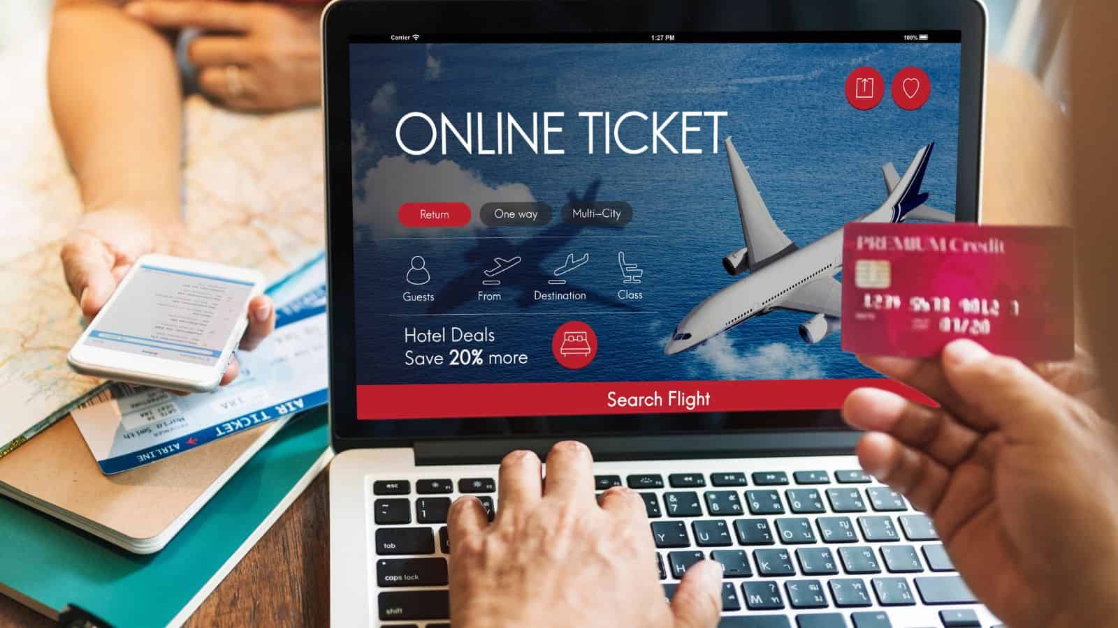 Person booking a flight online on a laptop with a credit card, passport, and phone visible on a table.