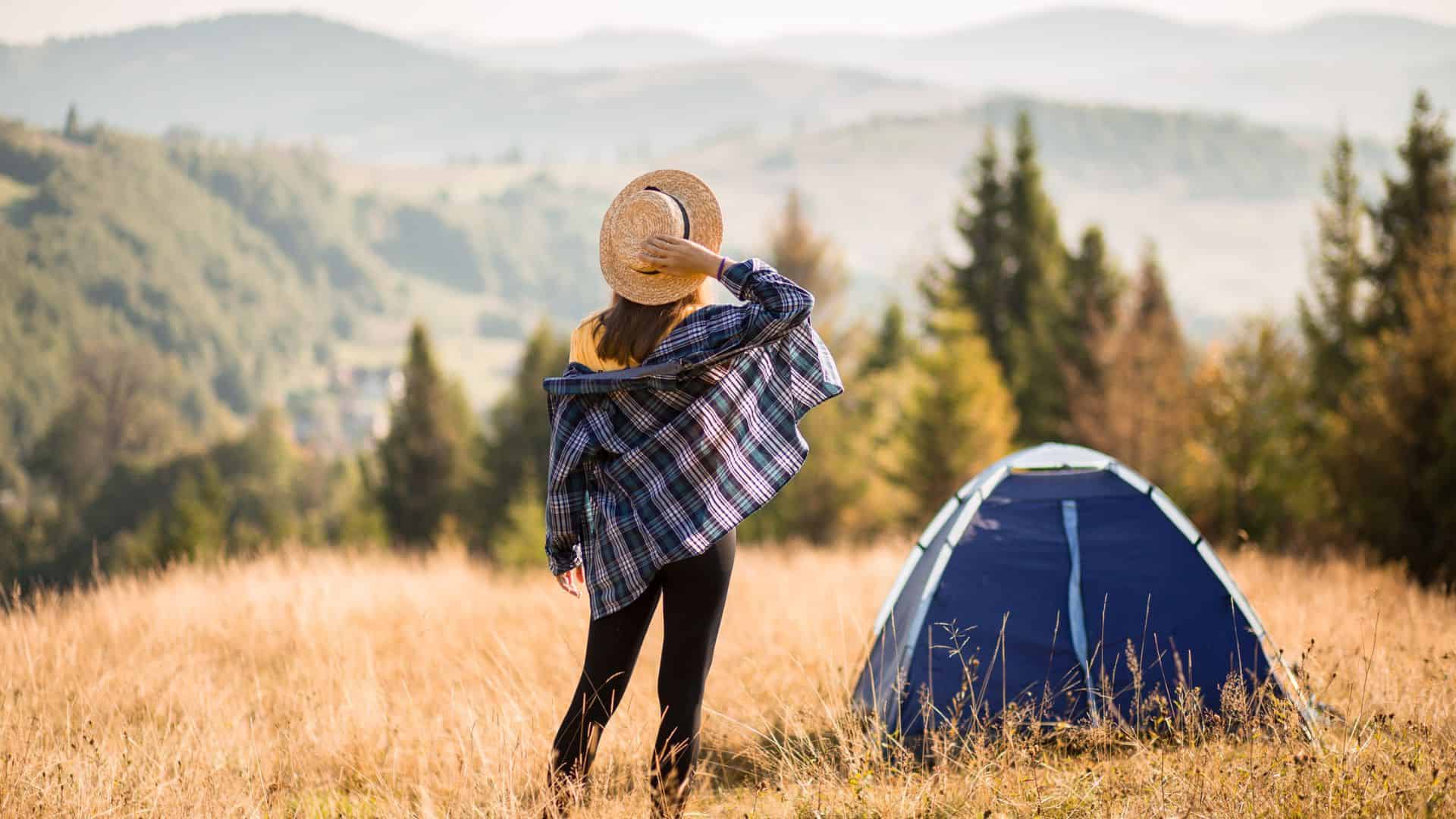 Woman in plaid shirt and hat stands in field near a blue tent, overlooking distant mountains and trees.