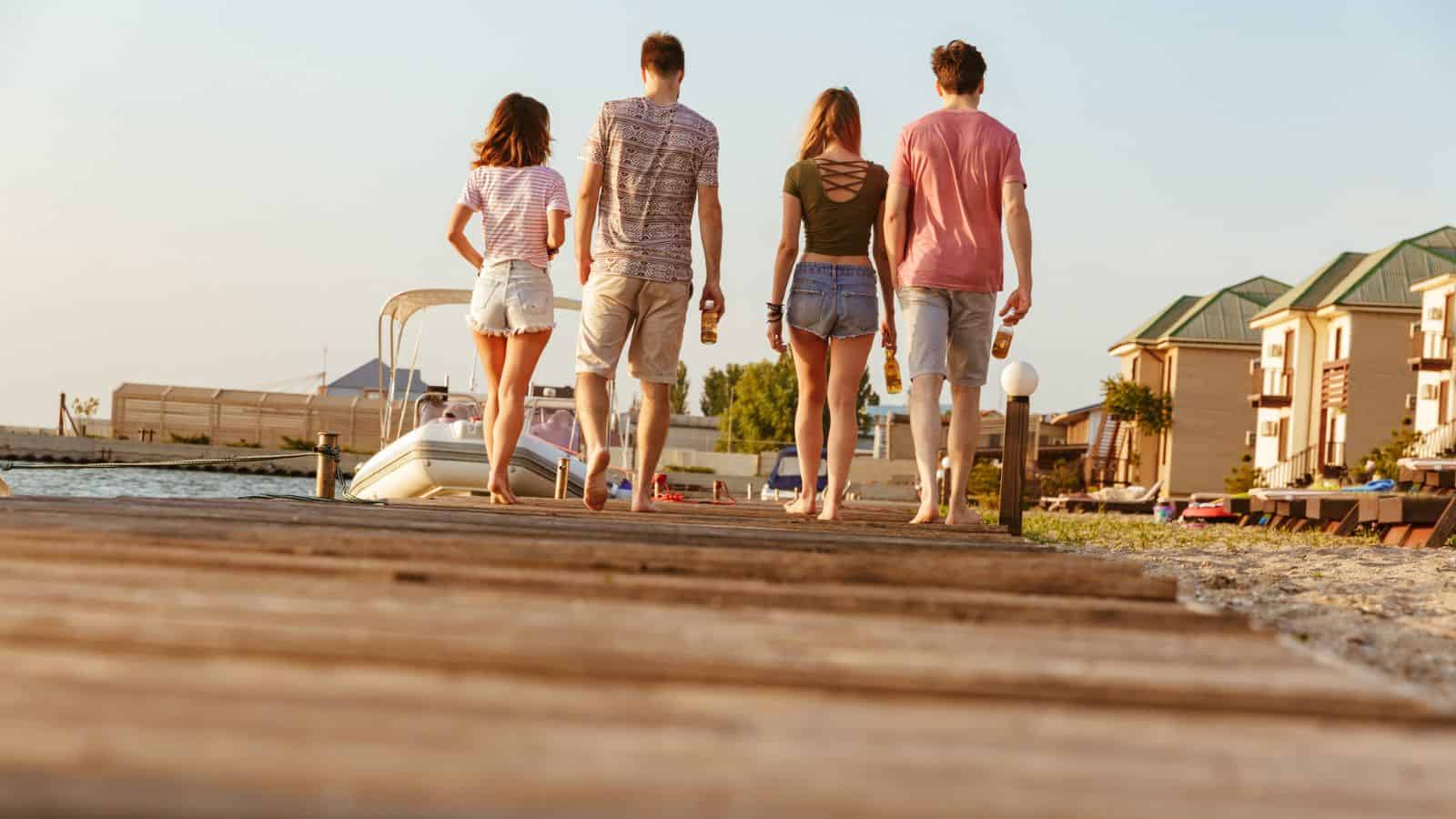 Four young adults walk barefoot on a wooden dock by the water, holding drinks on a sunny day.