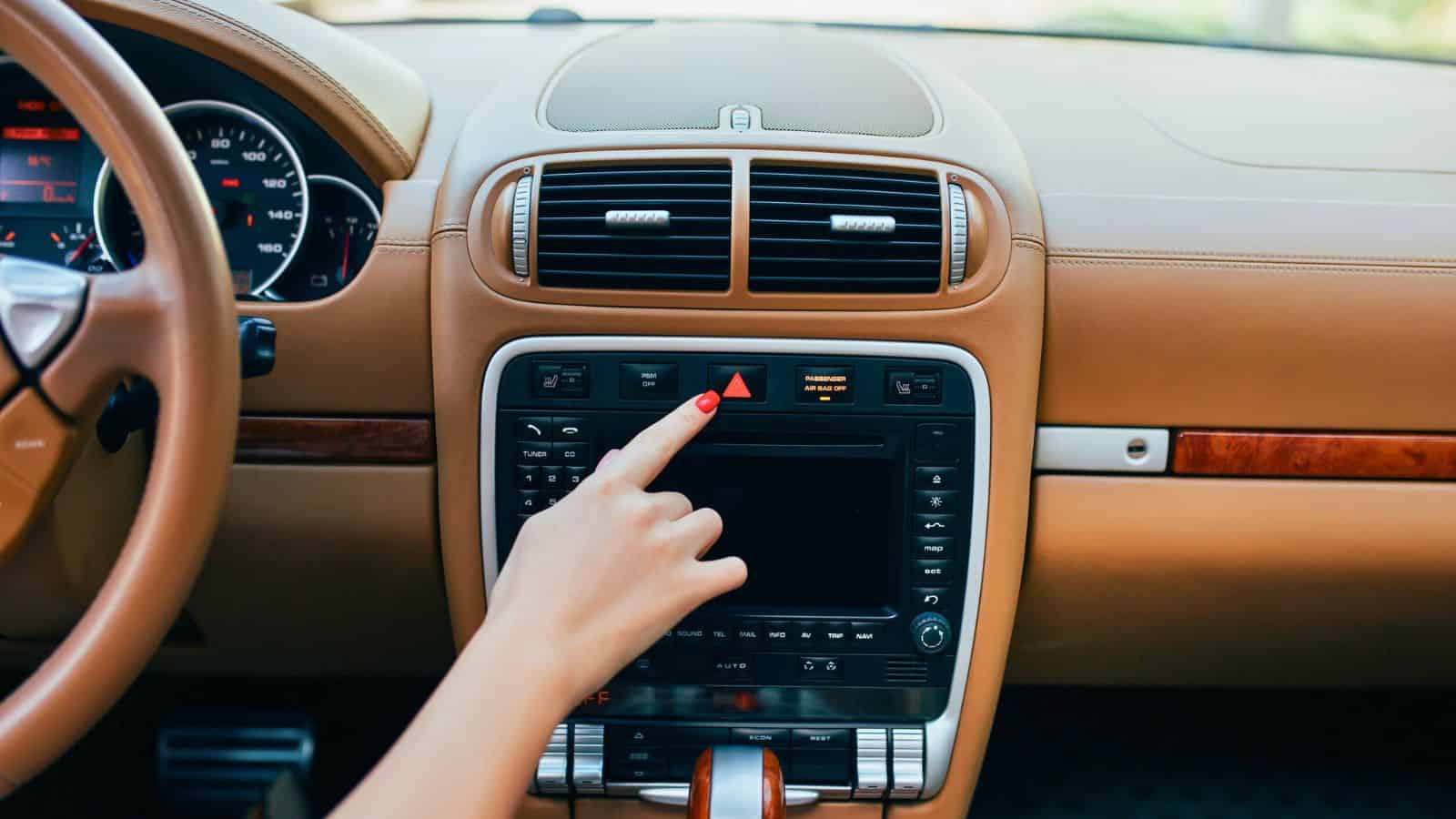 Hand pressing the hazard lights button on a car dashboard with a tan interior.
