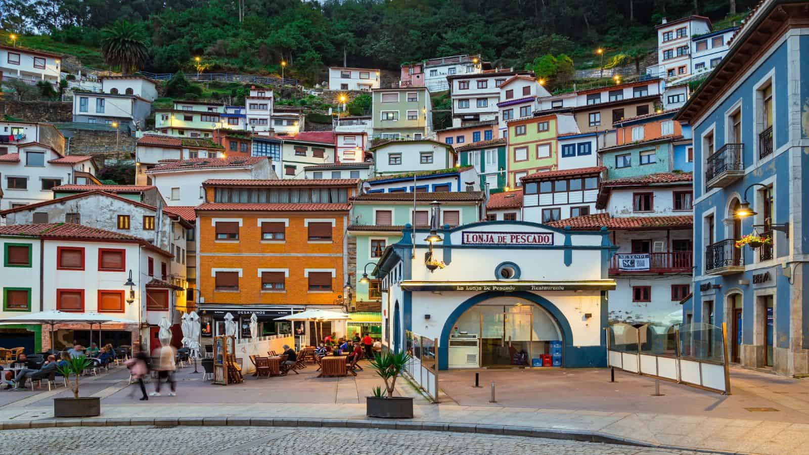 Colorful hillside houses and a small plaza with people sitting at outdoor cafés in a coastal village at dusk.