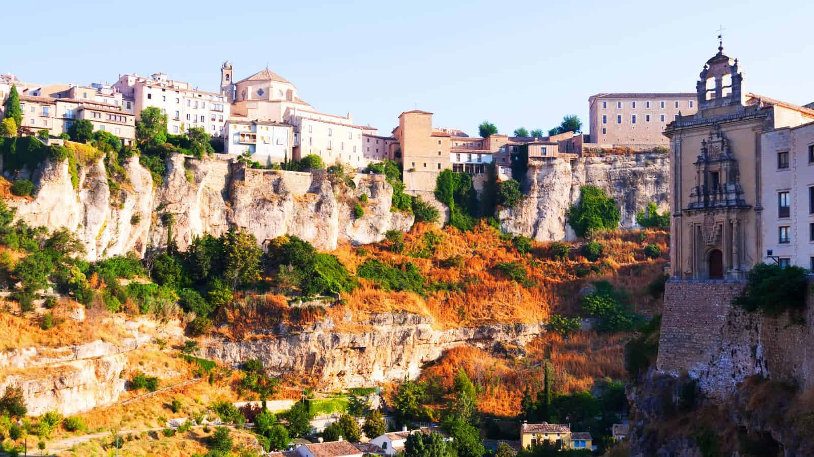 Historic buildings atop rugged cliffs with greenery and sunlit stone facades in a hillside town.