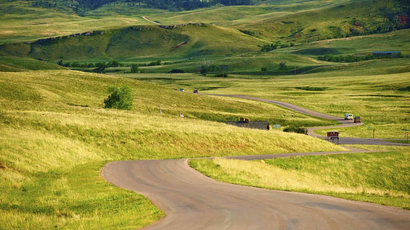 Winding road through rolling green hills under a clear sky, with scattered cars and lush grass&mdash;perfect for weekend getaways to hidden destinations.
