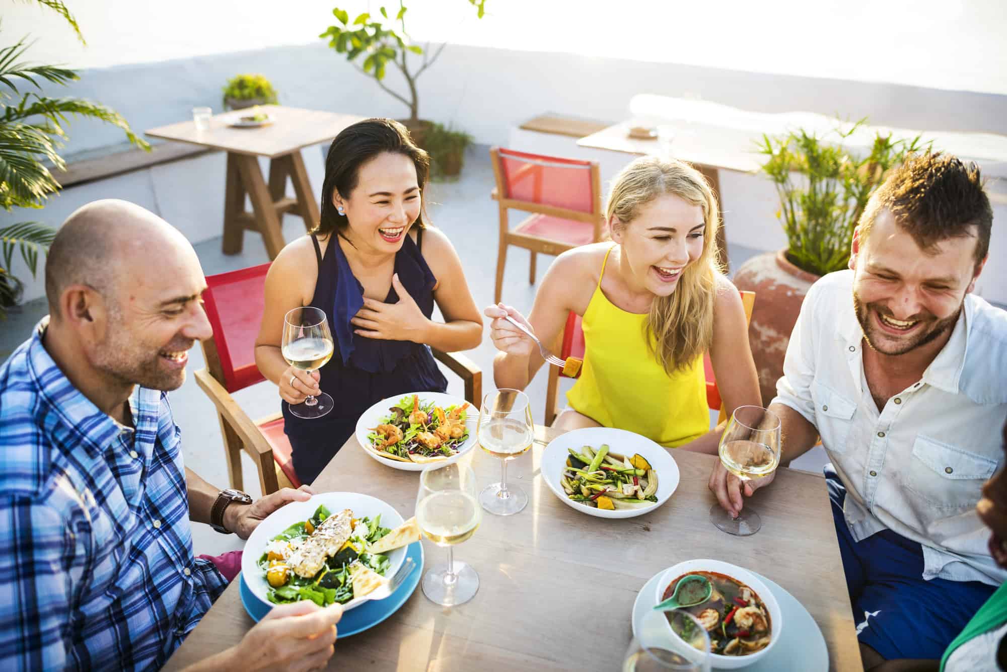 Four friends laughing and enjoying a meal together outside at one of the best restaurants in Barcelona, each with food and drinks at a sunny table.