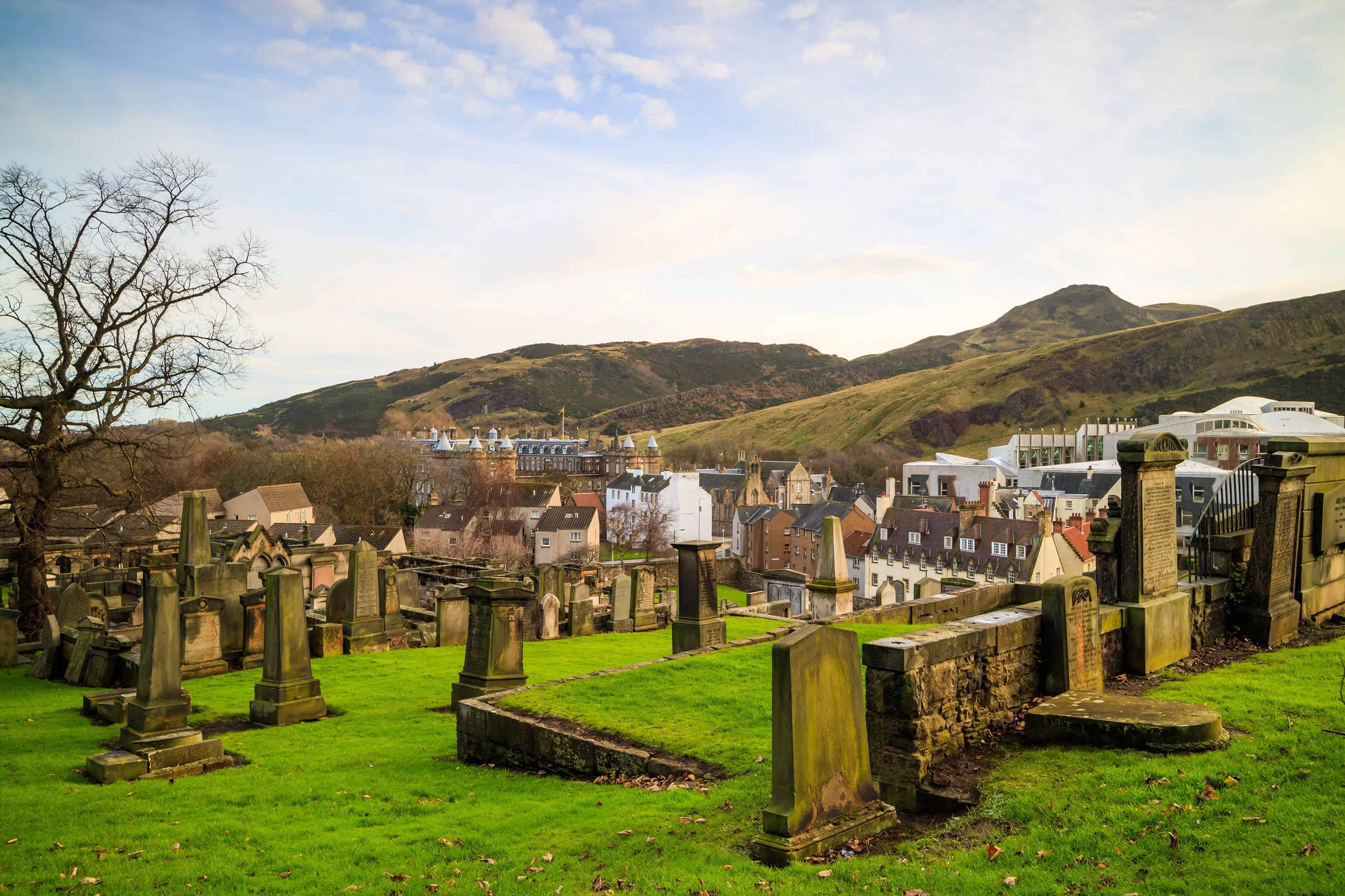 Old gravestones on a green hillside overlook a town with rolling hills and mountains in the background, evoking the mysterious charm often found in haunted places in Europe.