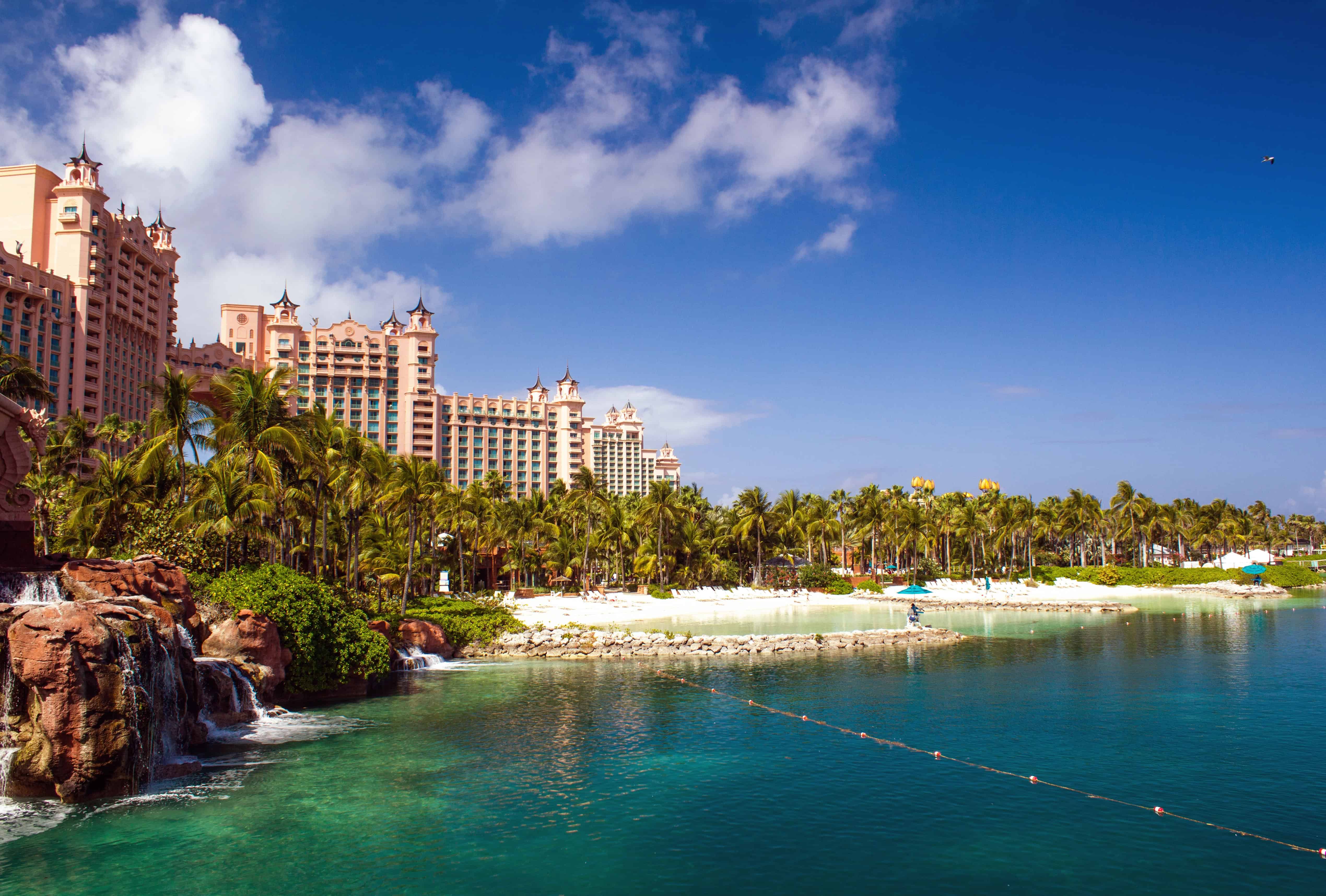A tropical resort with tall pink buildings, palm trees, waterfalls, and clear blue water under a bright sky—perfect for Bahamas cruise activities for families seeking fun and relaxation.
