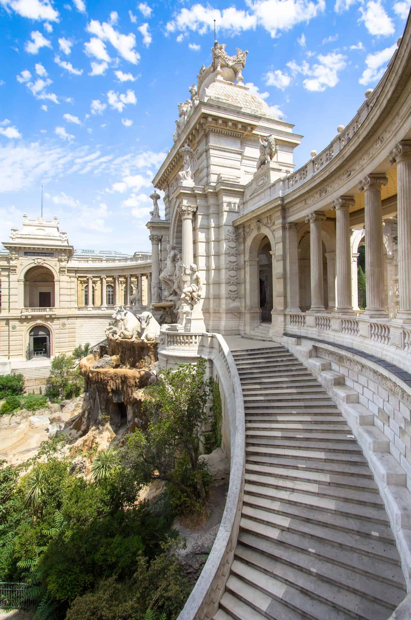 Curved stone staircase leads to a grand neoclassical building with columns, statues, and lush greenery—a must-see on any Marseille itinerary in France.