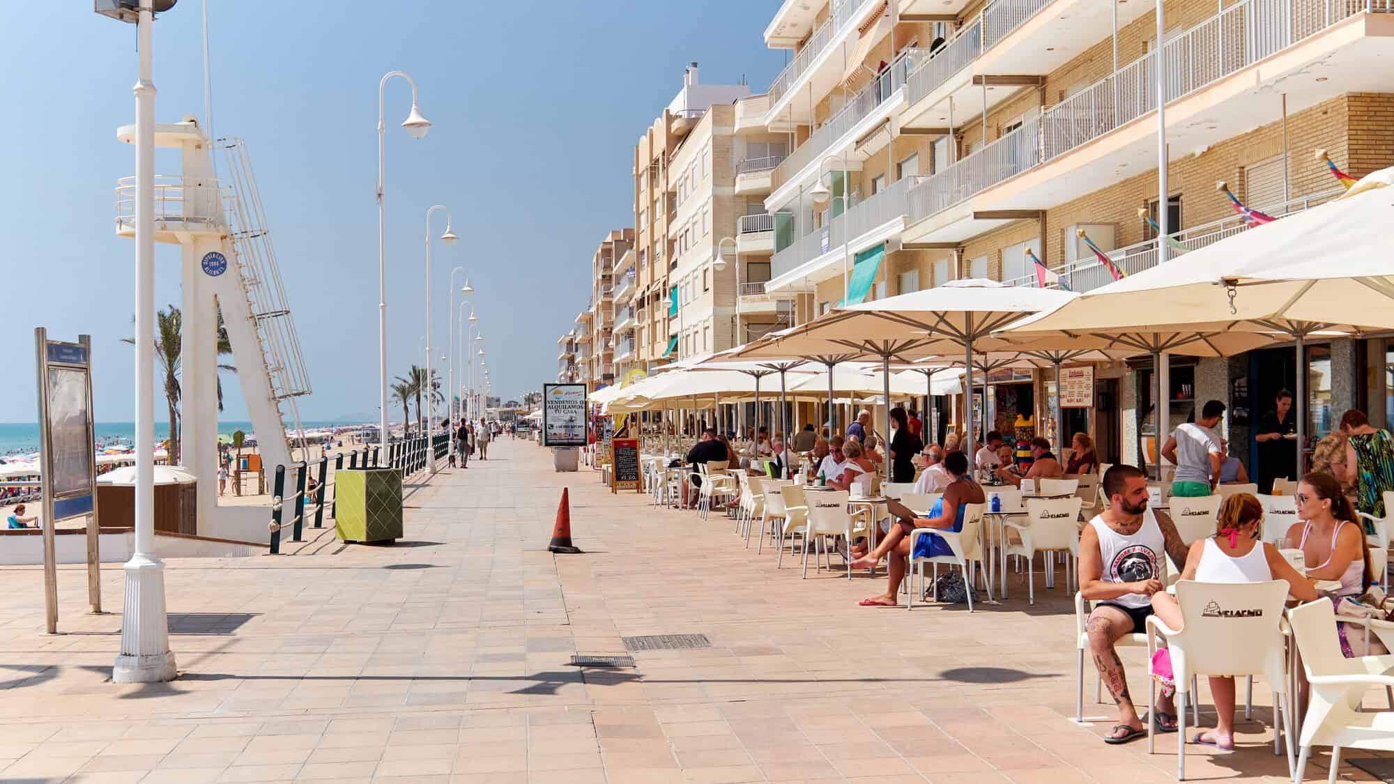 People dining at beachfront restaurants along a sunny beach promenade in Barcelona, with apartments and the sparkling sea in the background.
