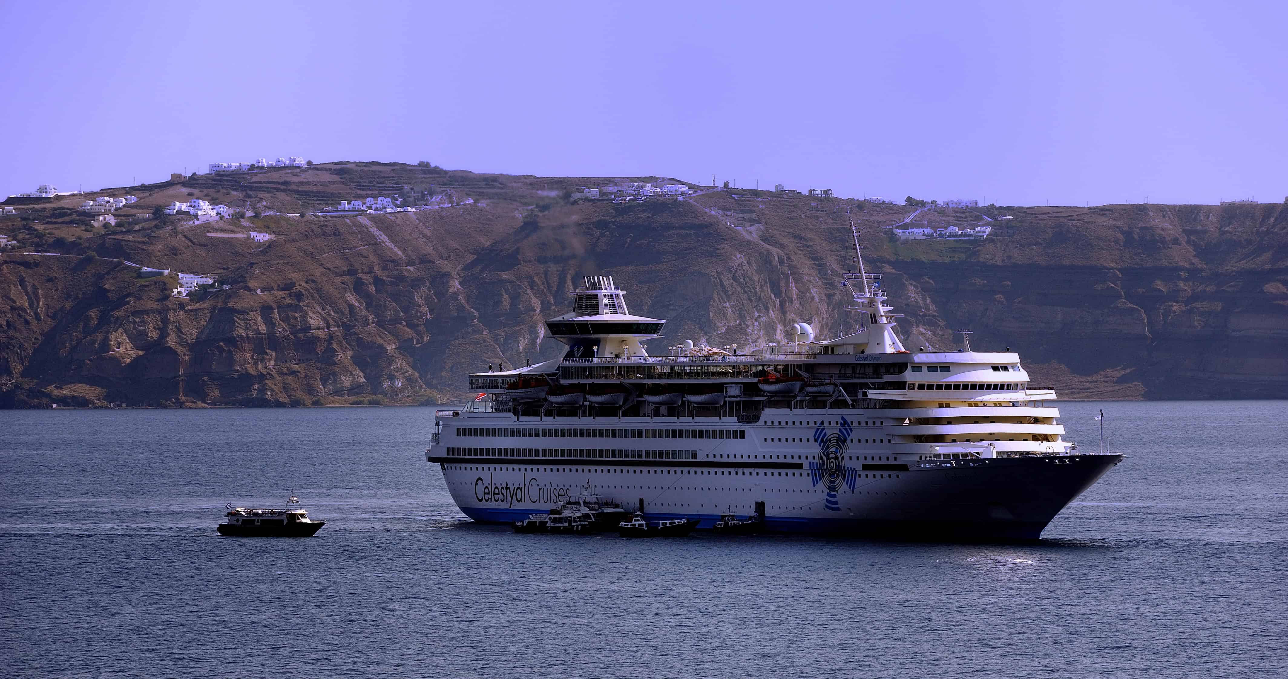 A large cruise ship sails near a rocky coastline with white buildings on the cliffs in the background, offering unforgettable cruises for adults seeking relaxation and stunning views.