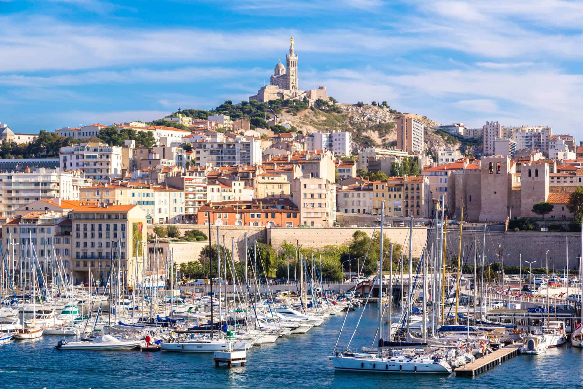 Boats docked in a marina with city buildings and a hilltop church in the background under a blue sky, capturing the charm of Marseille—a must-see stop on any France itinerary.