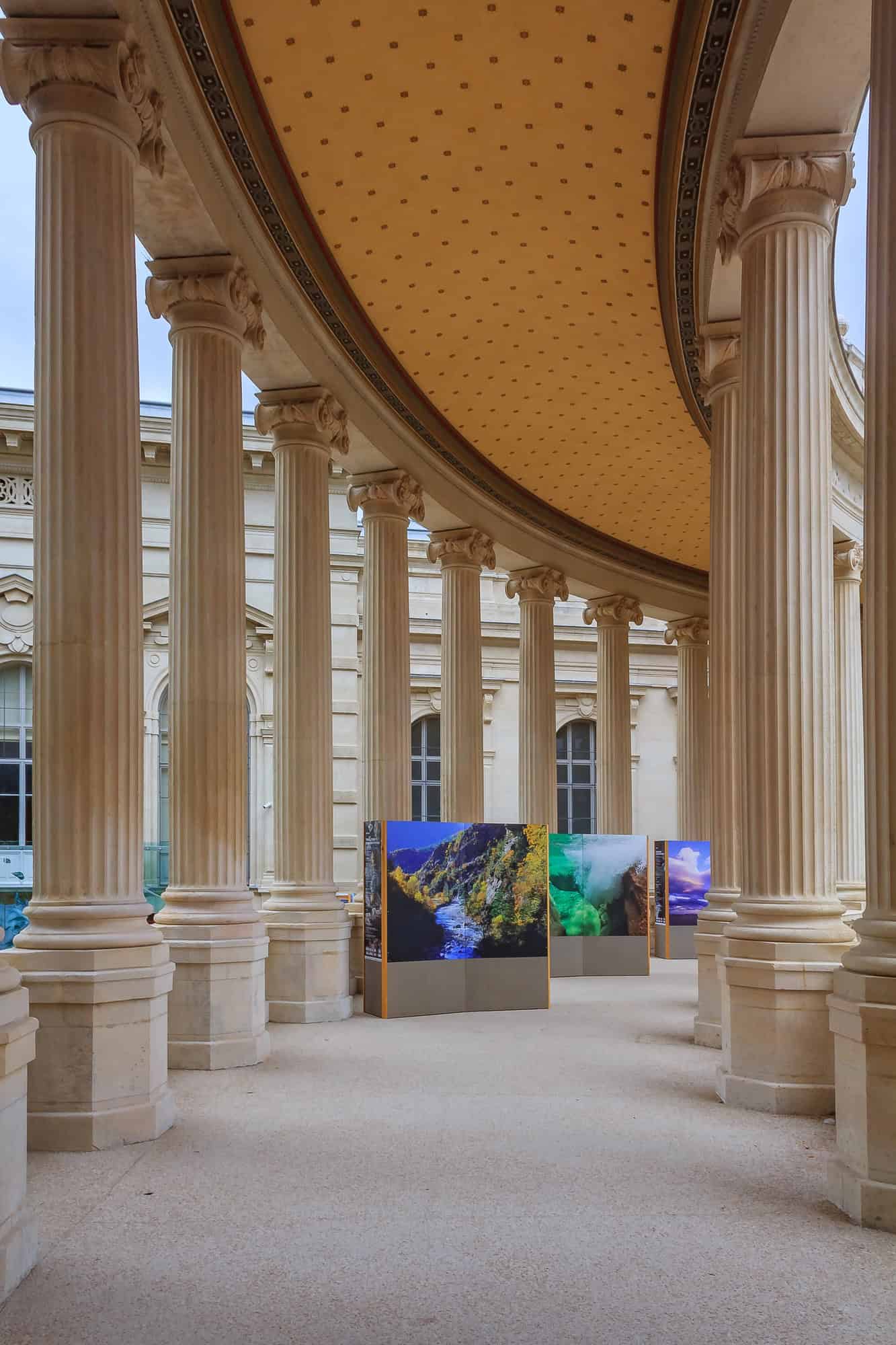 A colonnade with tall pillars in Marseille displays colorful landscape photographs on easels, making it a memorable stop on your France itinerary.