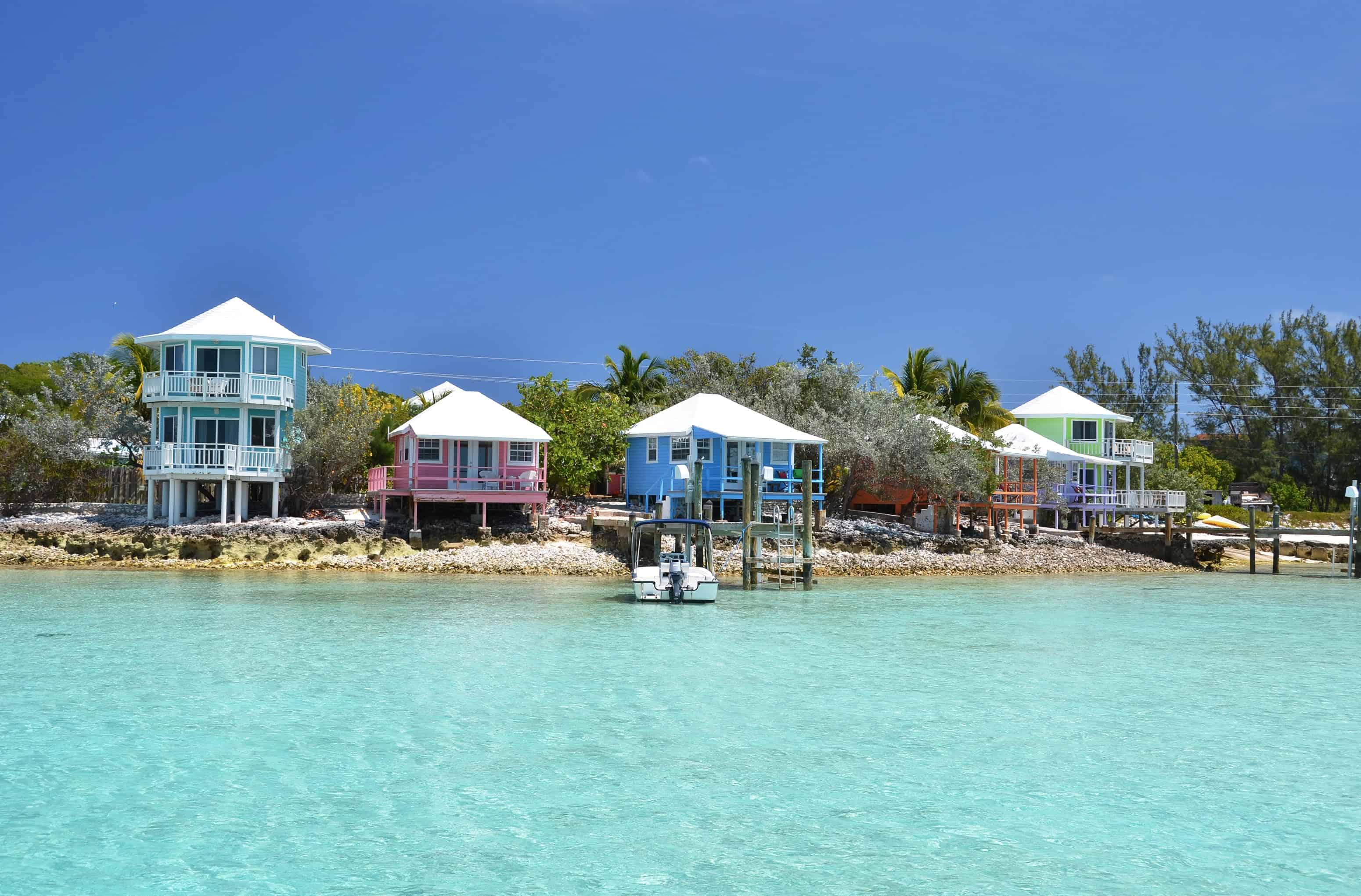 Colorful beachfront cottages with docks and a small boat on clear turquoise water under a sunny blue sky, perfect for enjoying Bahamas cruise activities for families.