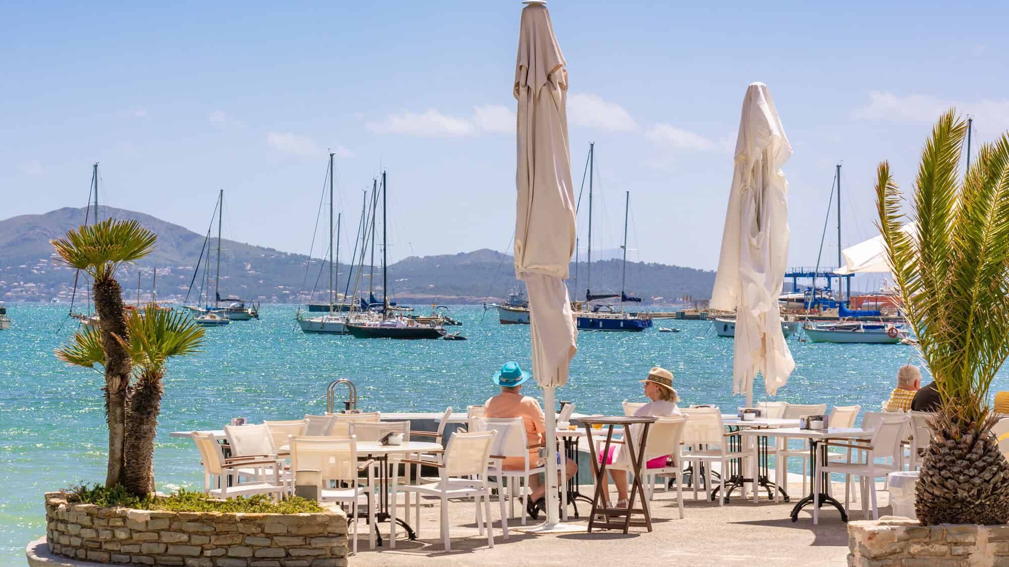 People enjoy Barcelona dining at a seaside café, with sailboats and mountains in the background under a sunny sky—one of the charming beachfront restaurants Barcelona has to offer.