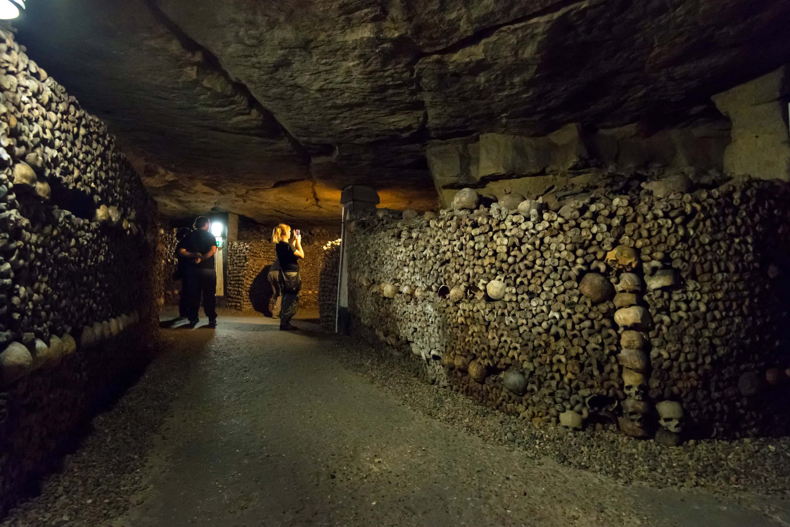 Two people walk in an underground tunnel lined with human skulls and bones in dim lighting—a chilling journey through one of the haunted places in Europe.