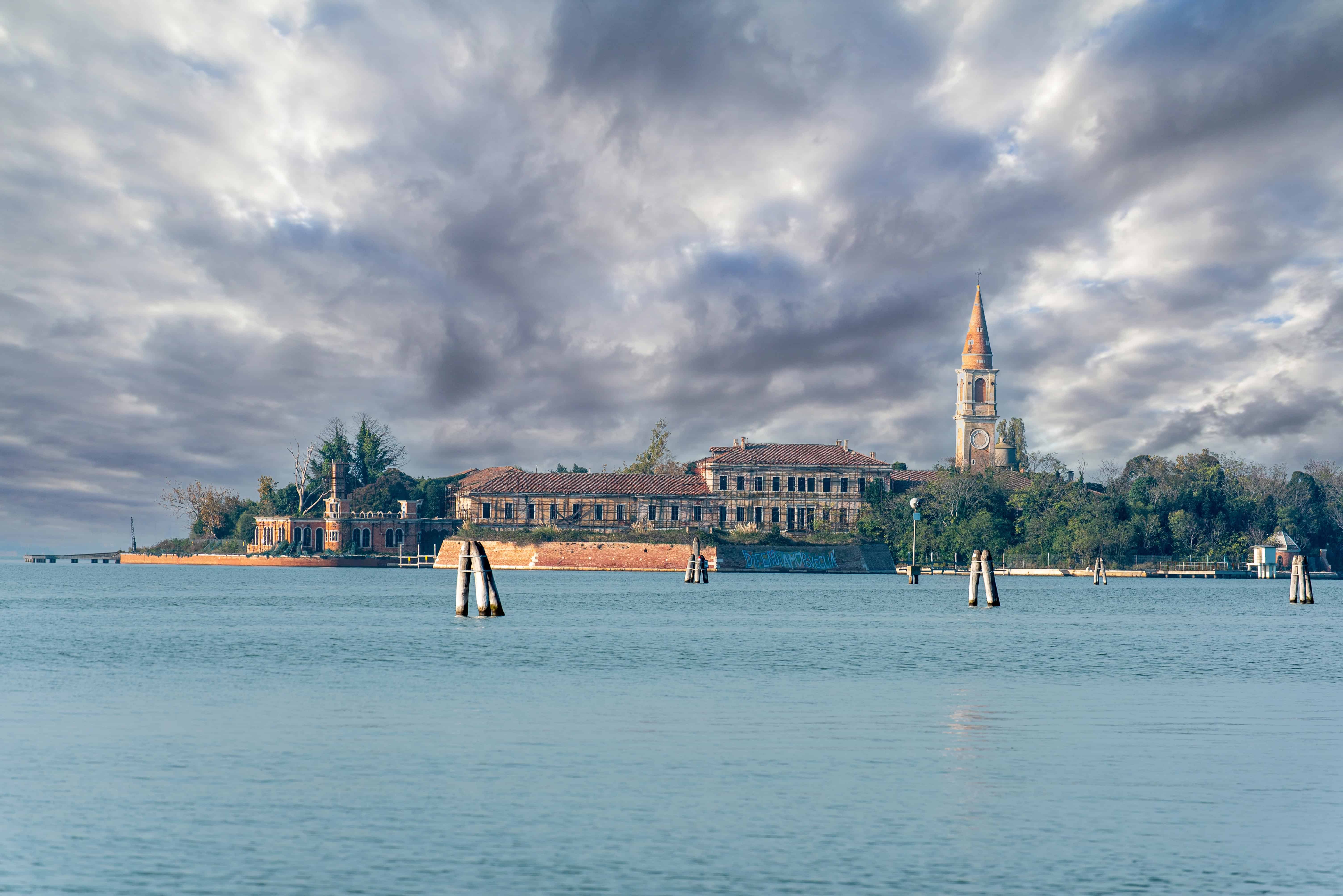 Island with old buildings, a tall bell tower, and cloudy sky, surrounded by calm water—an atmosphere reminiscent of haunted places in Europe.