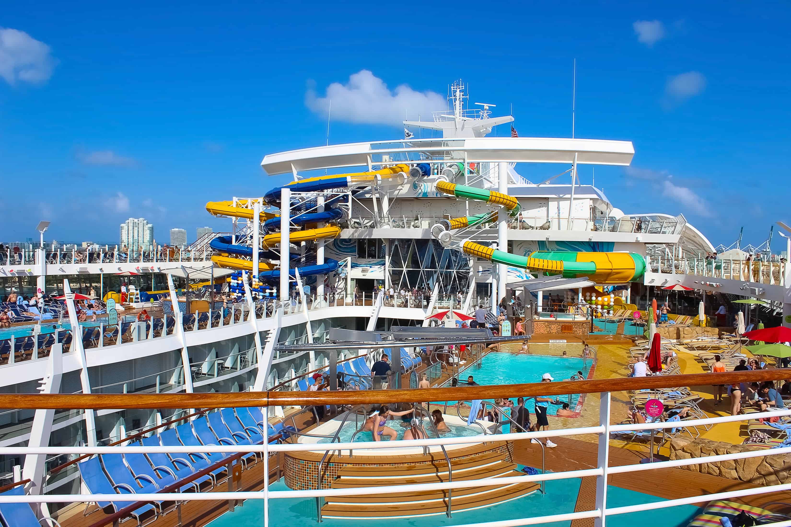 A cruise ship deck with pools, waterslides, lounge chairs, and people enjoying sunny weather—perfect for cruises for families looking to relax and have fun together.
