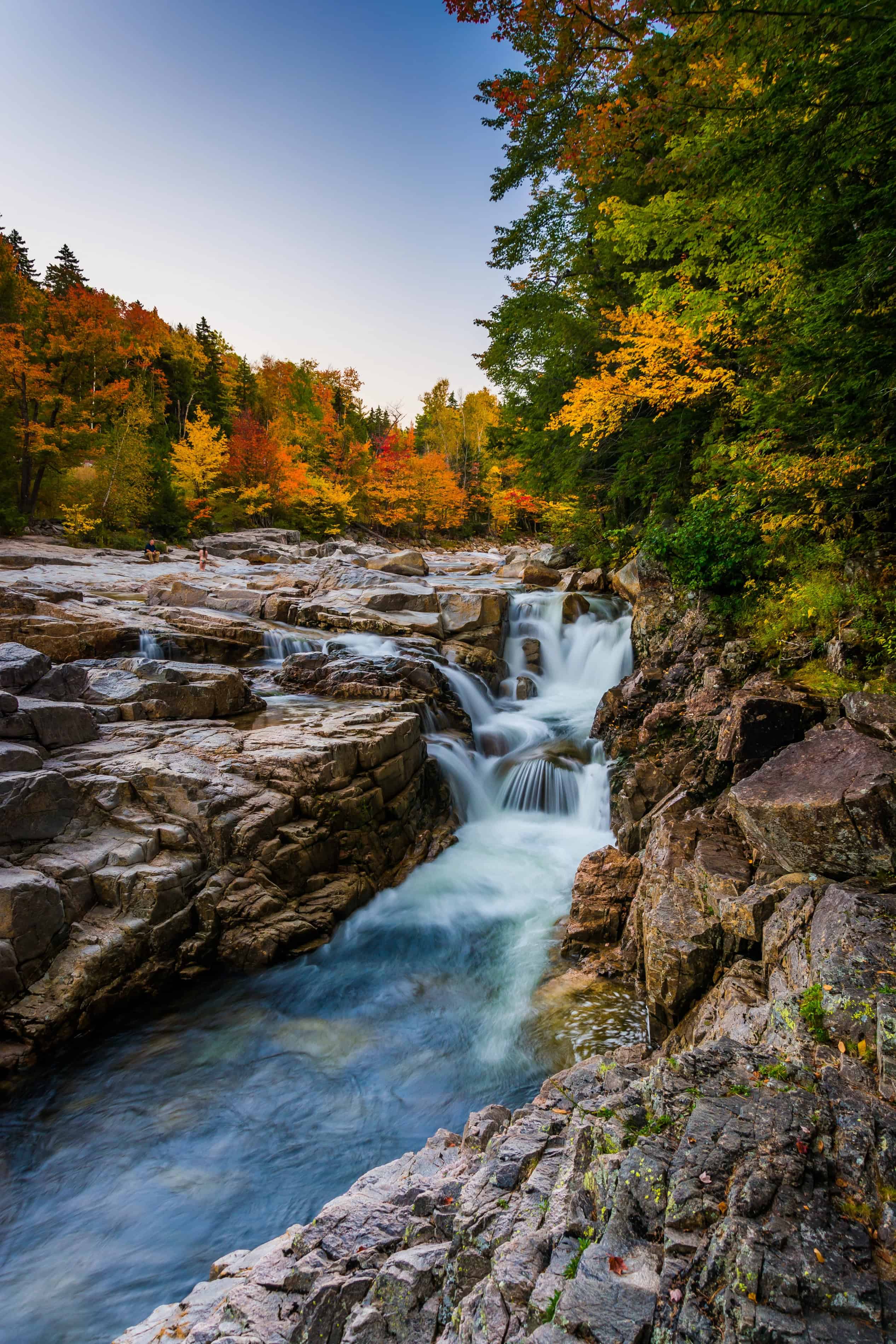 A small waterfall flows over rocks surrounded by the best fall foliage in USA, with colorful autumn trees under a clear sky.