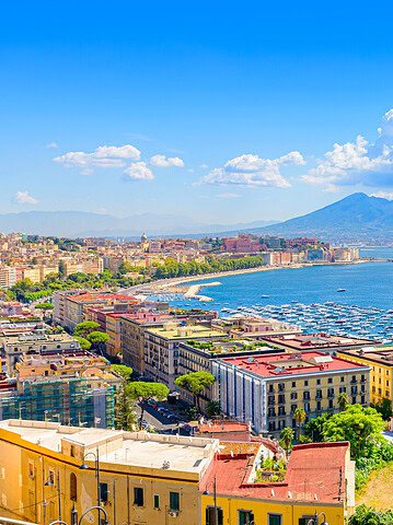 Colorful coastal city with boats in a bay, and a large volcano in the background under a blue sky—perfect for exploring Naples, Italy, and planning a 1 day itinerary to nearby Pompeii.