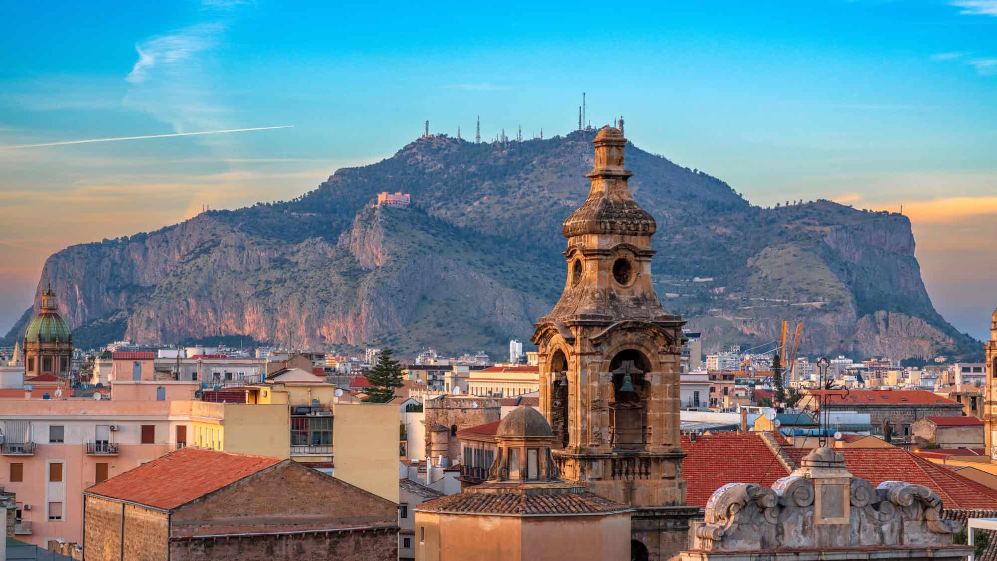 Rooftops and a historic church tower with a mountain in the background at sunset in Palermo, Sicily&mdash;a perfect scene to capture while exploring what to do in Palermo for a day.