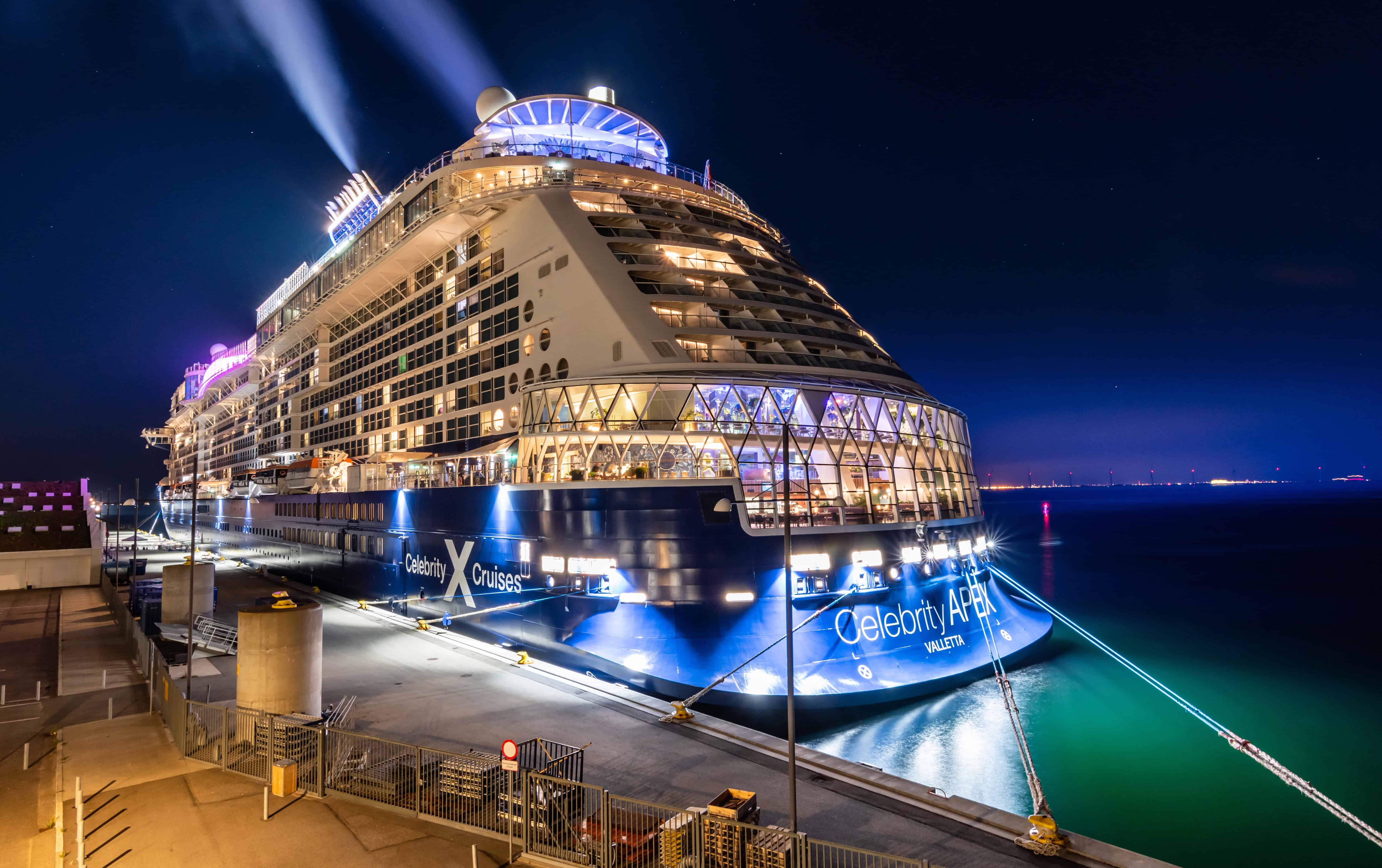 A brightly lit Celebrity Apex cruise ship, perfect for cruises for families, docked at night and reflecting on the water.