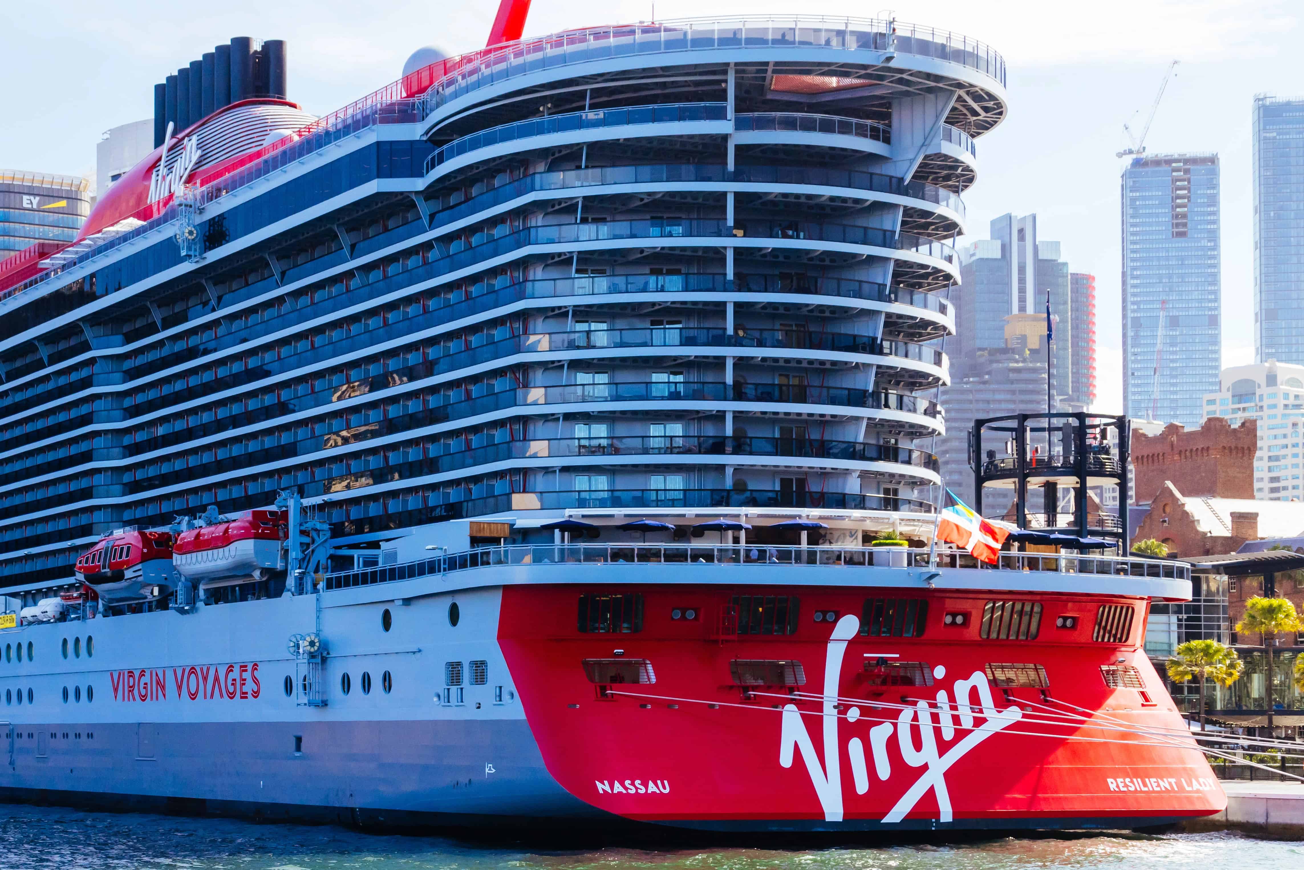 A large Virgin Voyages cruise ship docked in a city harbor with modern buildings in the background, showcasing an exciting option among cruise lines for adults seeking a stylish urban getaway.