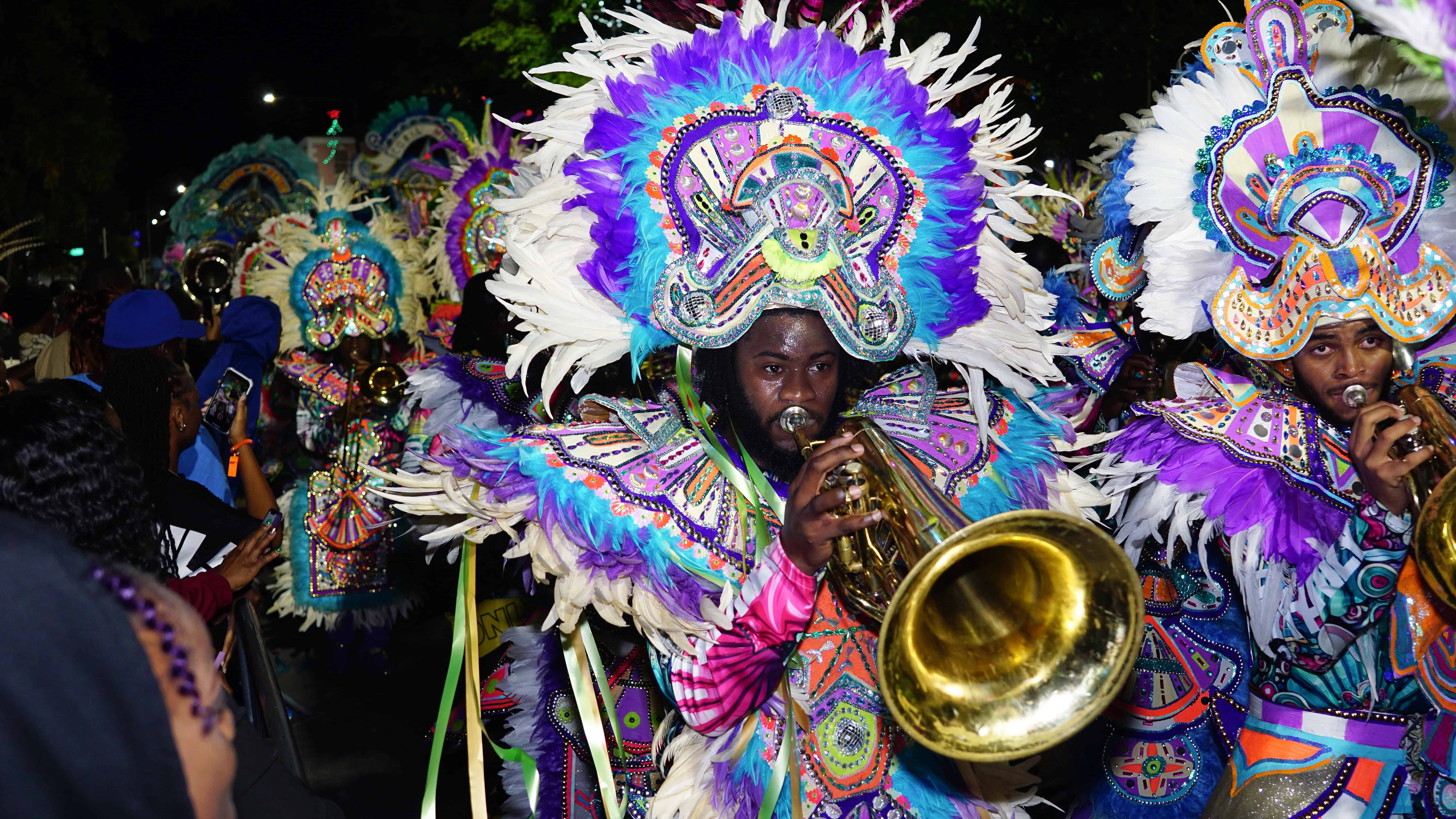 People in colorful feathered costumes play brass instruments during a vibrant street parade at night, showcasing festive Bahamas cruise activities for families.