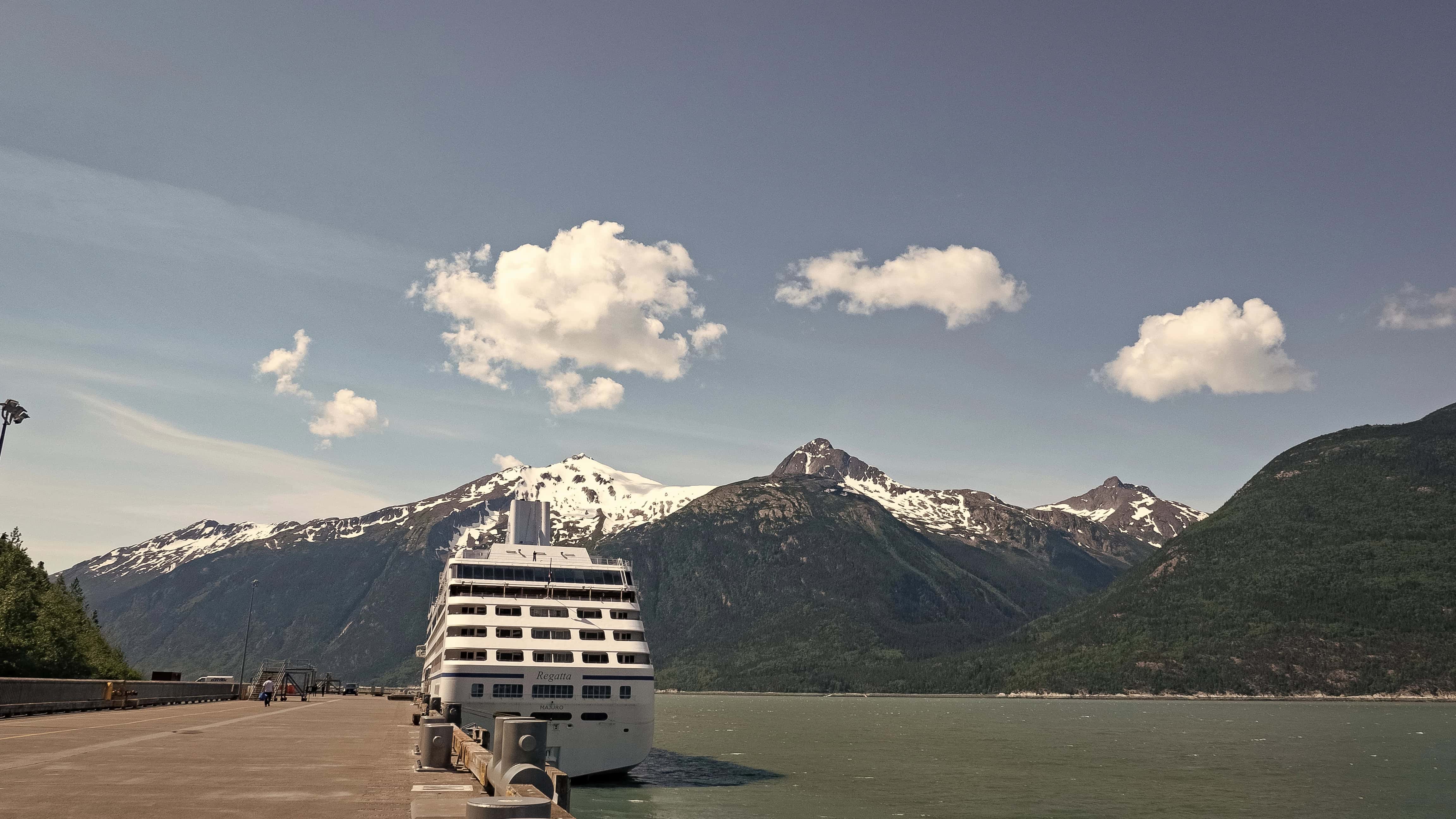 A cruise ship docked at a pier with snow-capped mountains and a cloudy sky in the background, offering an unforgettable experience for cruise lines for adults seeking scenic adventures.