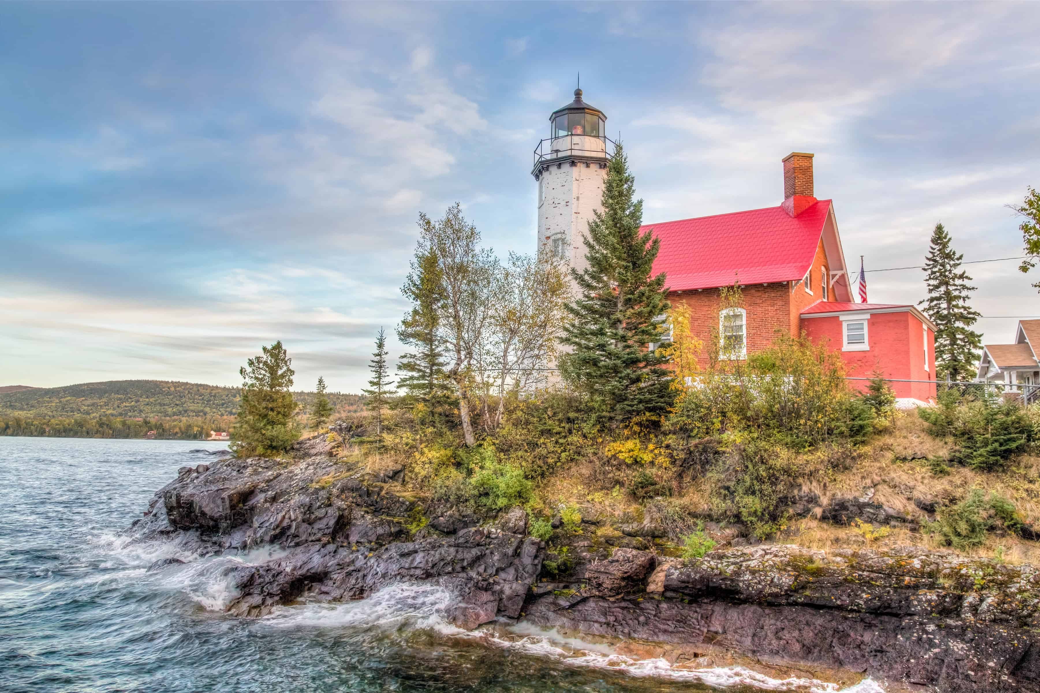 A lighthouse with a red roof stands on a rocky, tree-lined shore next to a calm body of water, surrounded by some of the best fall foliage in USA.