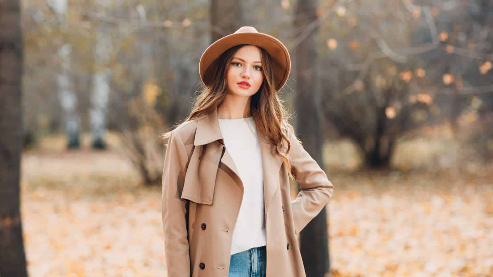 Woman in tan coat and hat standing outdoors among autumn trees with fallen leaves on the ground.