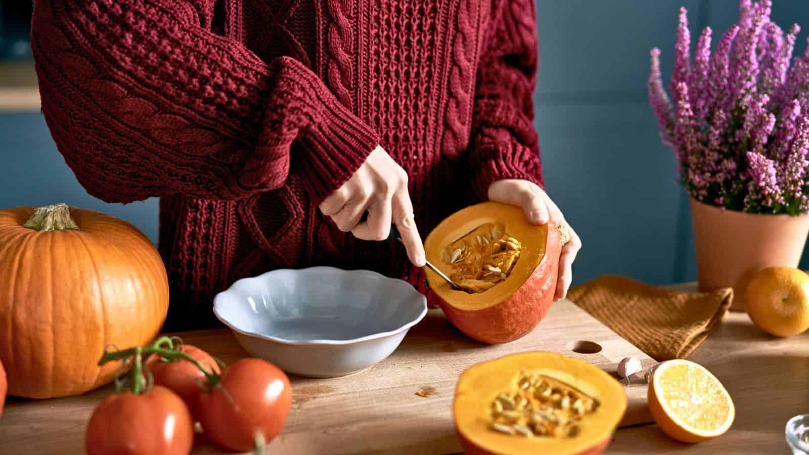 Person in a red sweater scooping seeds from a pumpkin at a kitchen table with tomatoes and flowers.