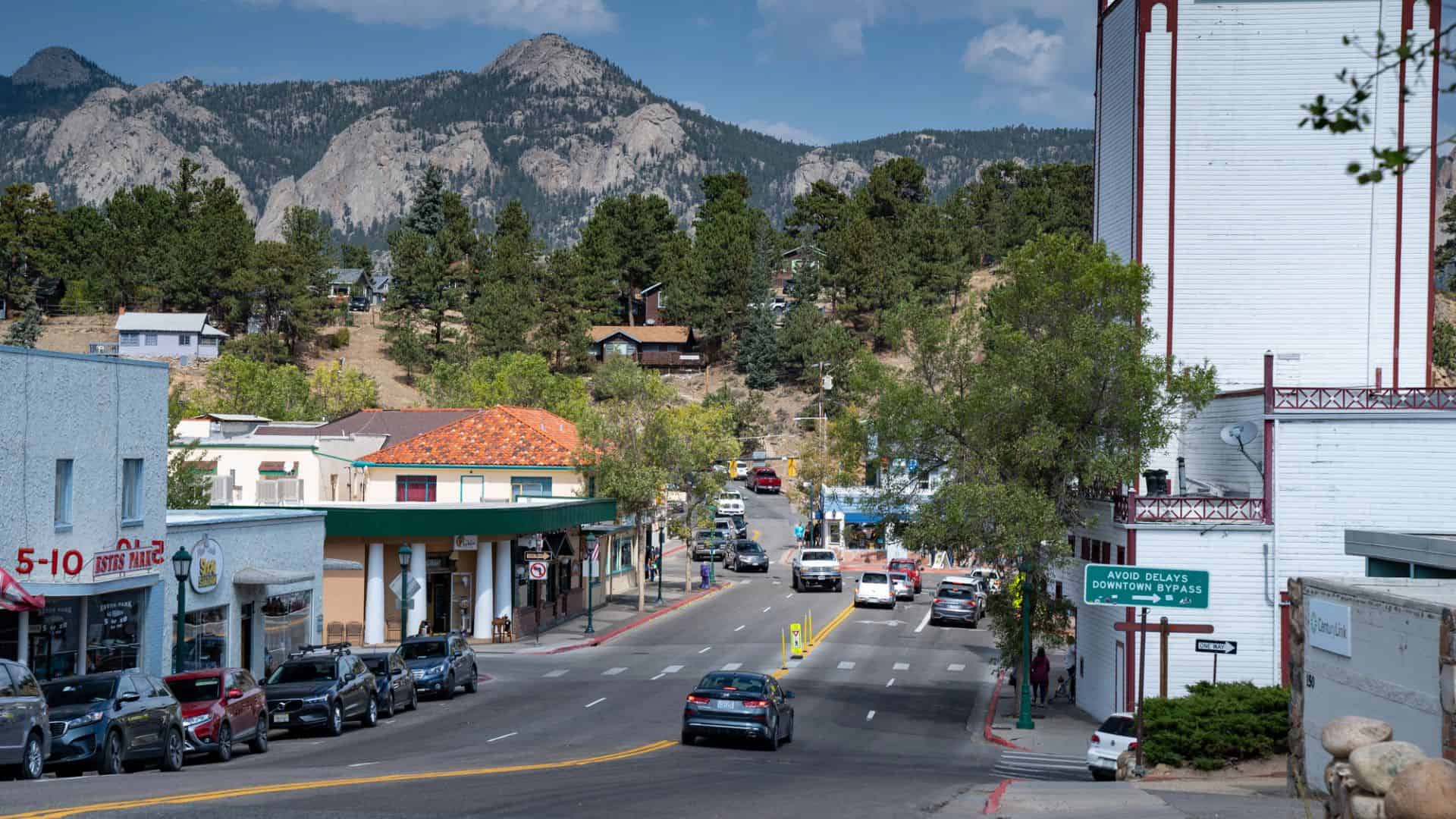 Small town street with cars, shops, and trees, with mountains in the background under a blue sky.