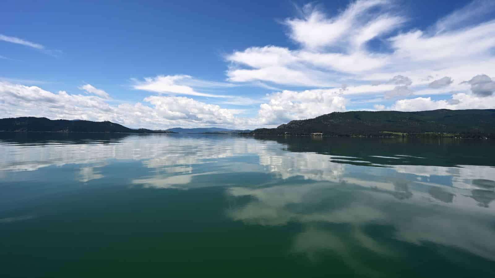 Calm lake with blue sky, white clouds, and distant green hills reflected in the water.