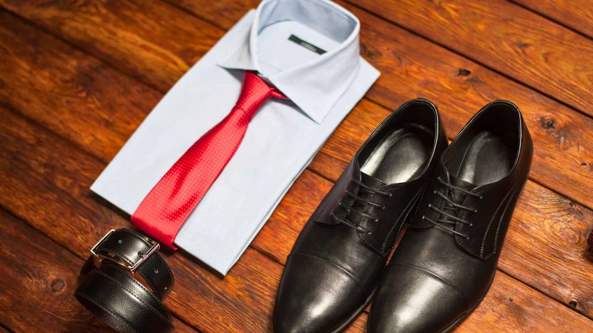 Folded blue shirt with red tie, black dress shoes, and a black belt on a wooden surface.