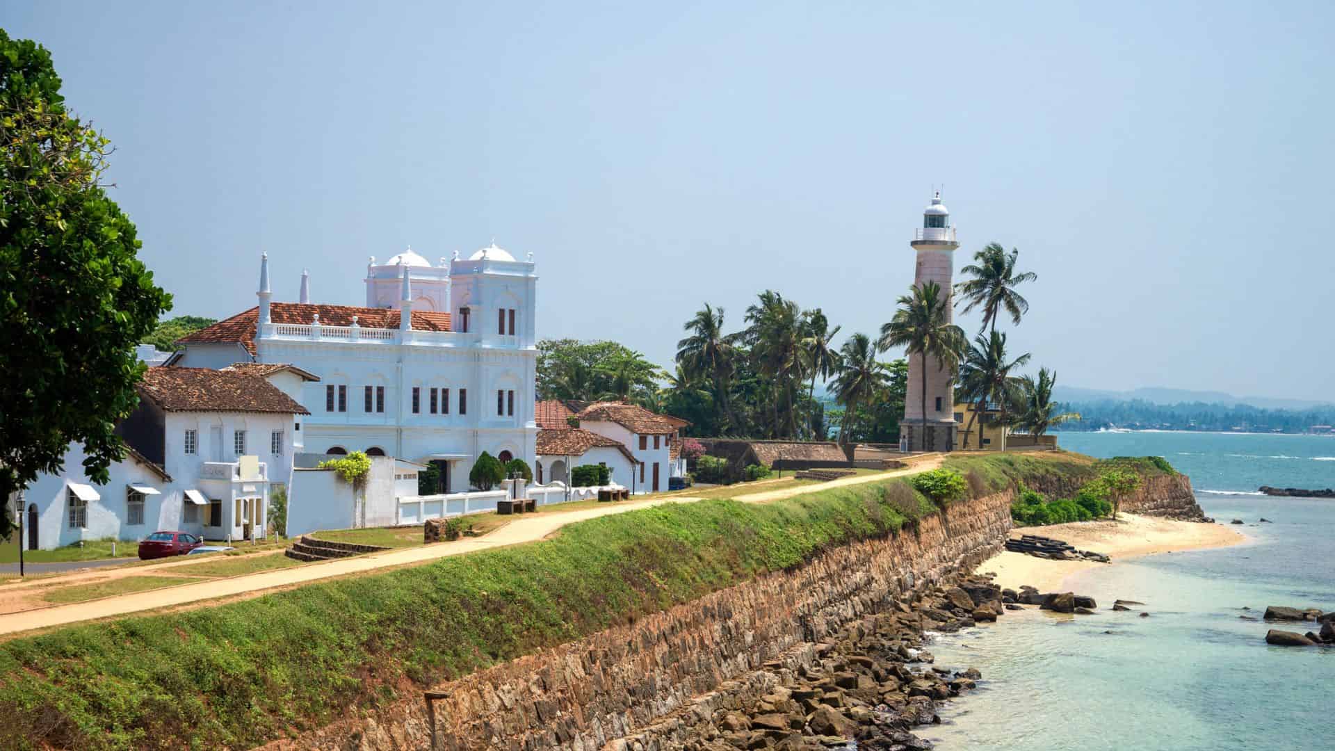 White colonial buildings and a lighthouse stand by the ocean, lined with palm trees and a stone wall in Galle, Sri Lanka.