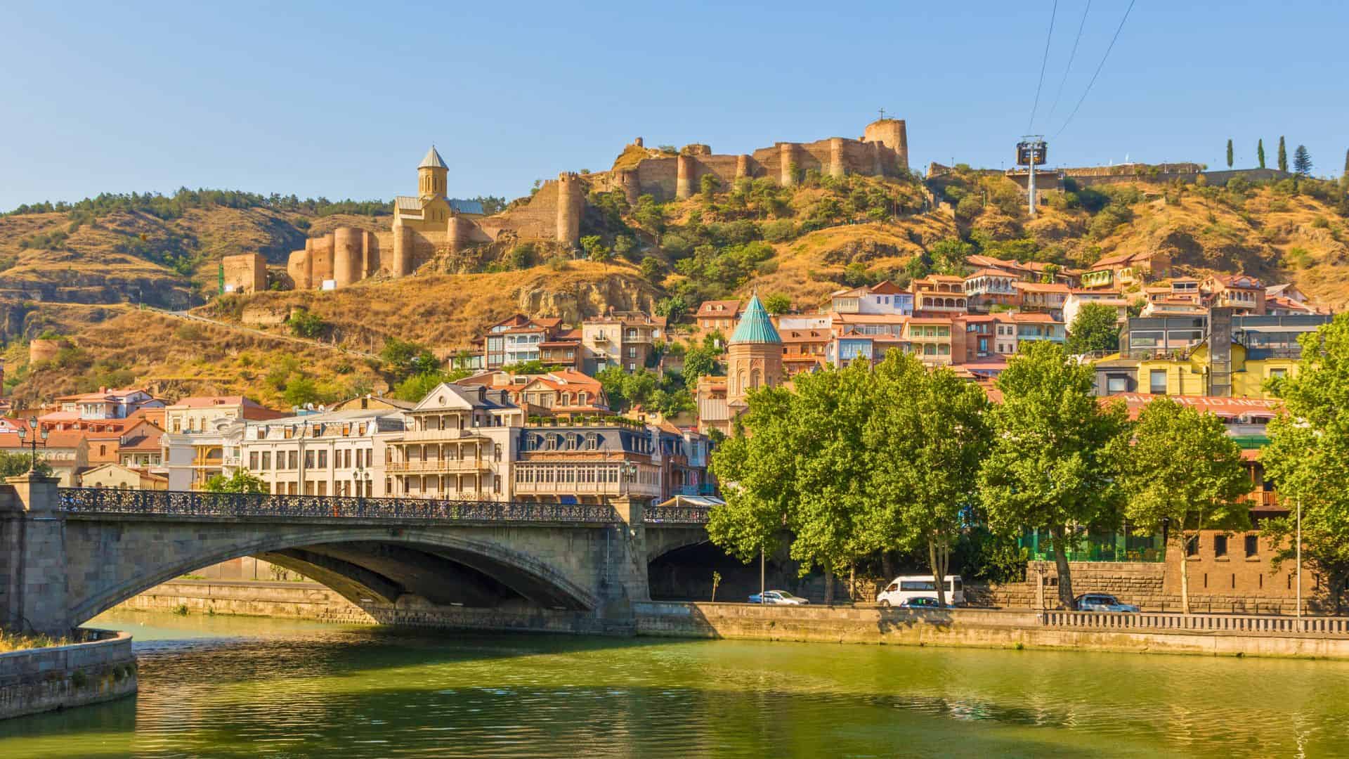 Historic fortress on a hill above colorful buildings and a bridge over a river in Tbilisi, Georgia.