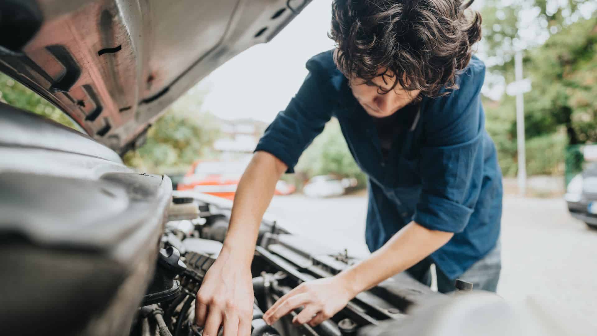 Person with curly hair checks under the hood of a car parked outdoors on a sunny day.